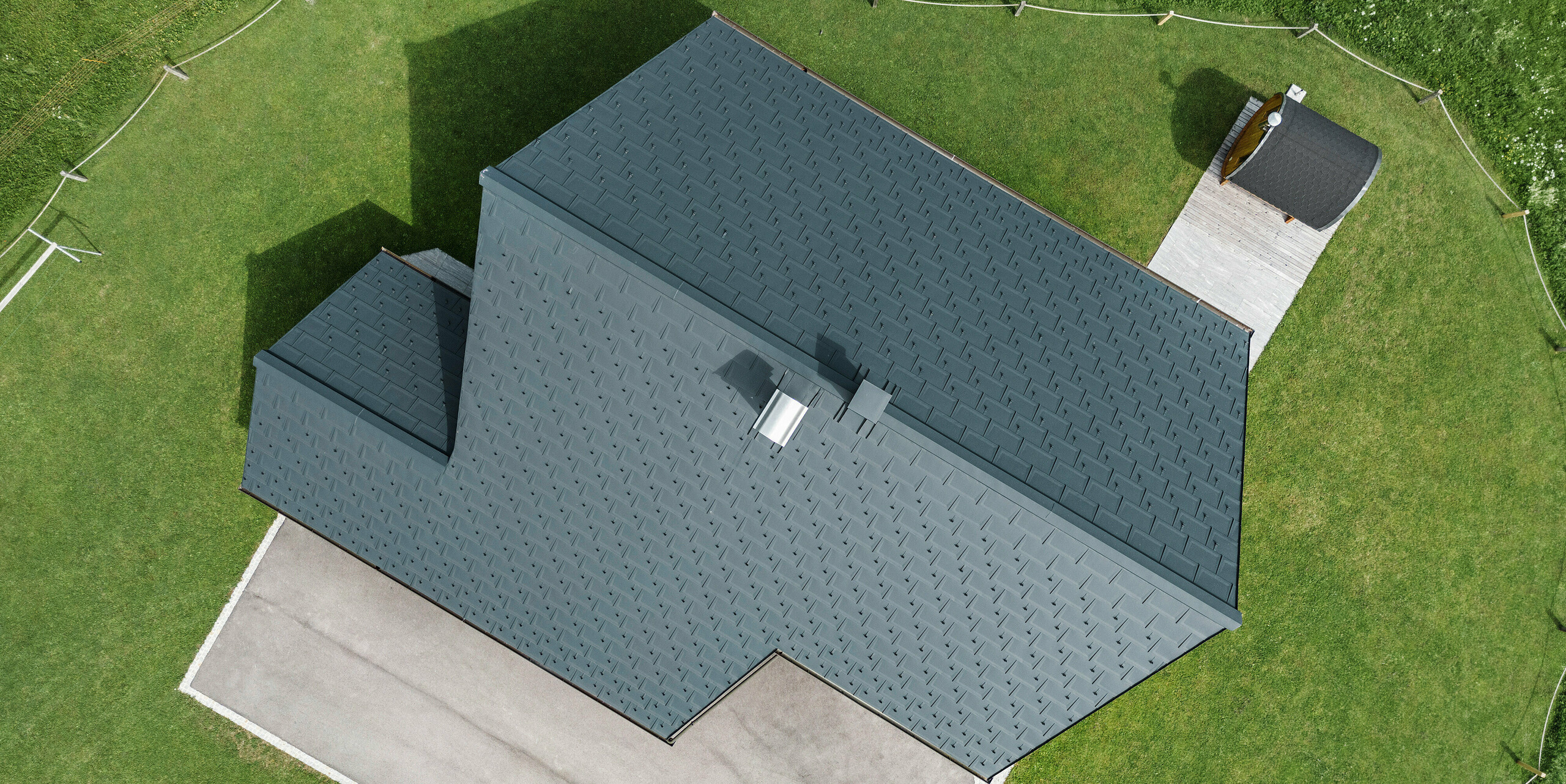 Vogelperspektive auf ein Blockhaus in Gosau, Oberösterreich, mit einem präzise verlegten PREFA Aluminiumdach aus Dachplatten R.16 in P.10 Anthrazit. Das Bild demonstriert das robuste Dachsystem und geometrische Eleganz. Die Dachplatten R.16 verleihen dem traditionell österreichischen Holzhaus moderne Konturen, während die umliegenden grünen Grasflächen und die klar definierten Wege die saubere und gepflegte Landschaftsgestaltung hervorheben.