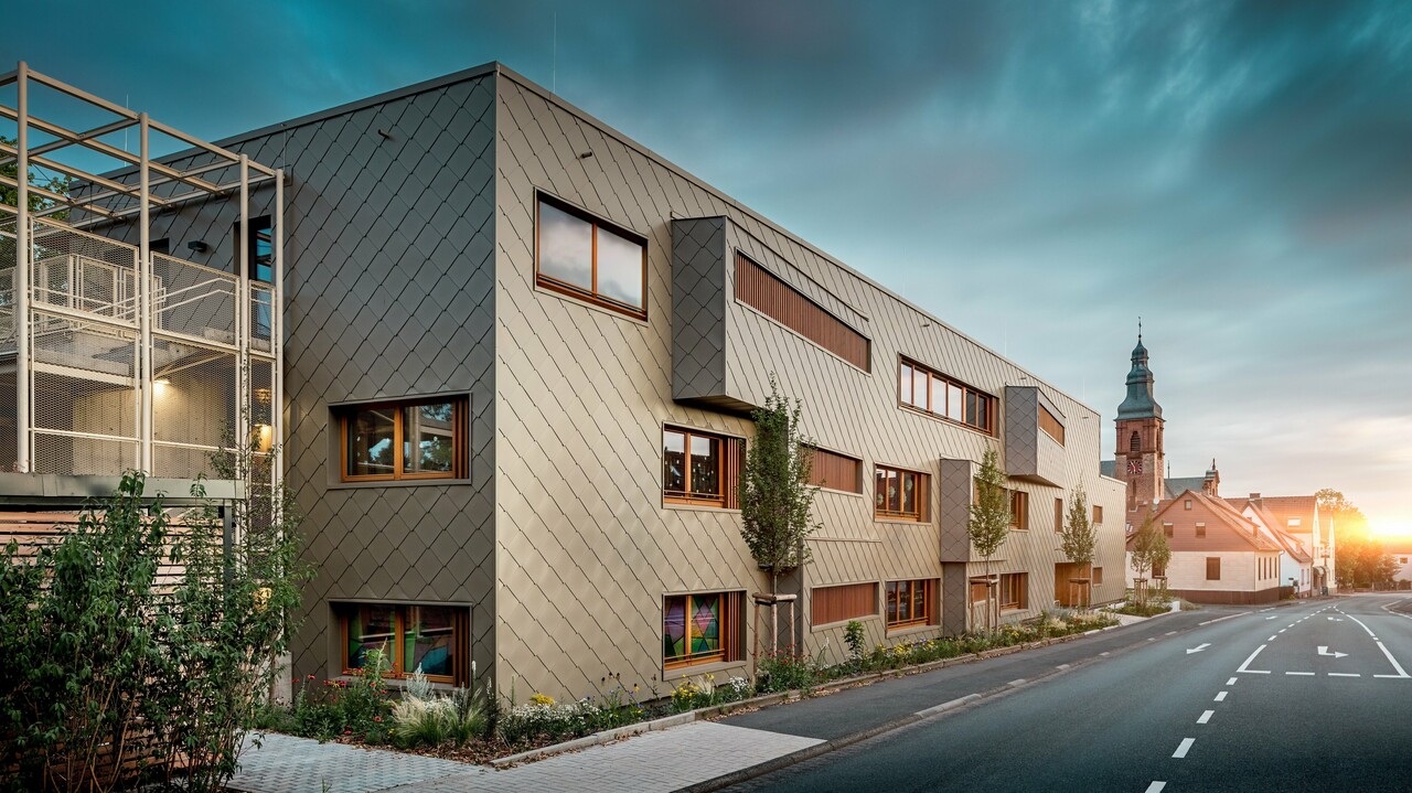 Im sanften Licht des Sonnenuntergangs zeigt sich die Kindertagesstätte in Alzenau von ihrer stimmungsvollsten Seite. Die Fassade aus bronzefarbenen PREFA Wandrauten 44 × 44 reflektiert das Abendlicht und betont die hochwertige, strukturierte Oberfläche des modernen Hybridgebäudes. Die klar gegliederte Architektur mit versetzten Fensterbändern und vertikalen Holzlamellen steht in spannendem Kontrast zur historischen Kirche im Hintergrund. Links im Bild sorgt ein filigranes Metallgerüst für leichte Transparenz beim Außentreppenhaus. Der gepflegte Grünstreifen entlang des Gebäudes unterstreicht den ökologischen Anspruch. Dieses Bild verbindet eindrucksvoll moderne Baukultur, nachhaltige PREFA Aluminiumtechnologie und harmonische Einbettung in das historische Stadtbild von Alzenau.