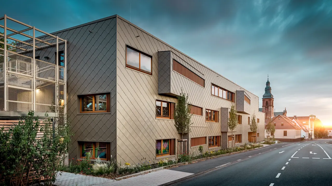 Im sanften Licht des Sonnenuntergangs zeigt sich die Kindertagesstätte in Alzenau von ihrer stimmungsvollsten Seite. Die Fassade aus bronzefarbenen PREFA Wandrauten 44 × 44 reflektiert das Abendlicht und betont die hochwertige, strukturierte Oberfläche des modernen Hybridgebäudes. Die klar gegliederte Architektur mit versetzten Fensterbändern und vertikalen Holzlamellen steht in spannendem Kontrast zur historischen Kirche im Hintergrund. Links im Bild sorgt ein filigranes Metallgerüst für leichte Transparenz beim Außentreppenhaus. Der gepflegte Grünstreifen entlang des Gebäudes unterstreicht den ökologischen Anspruch. Dieses Bild verbindet eindrucksvoll moderne Baukultur, nachhaltige PREFA Aluminiumtechnologie und harmonische Einbettung in das historische Stadtbild von Alzenau.