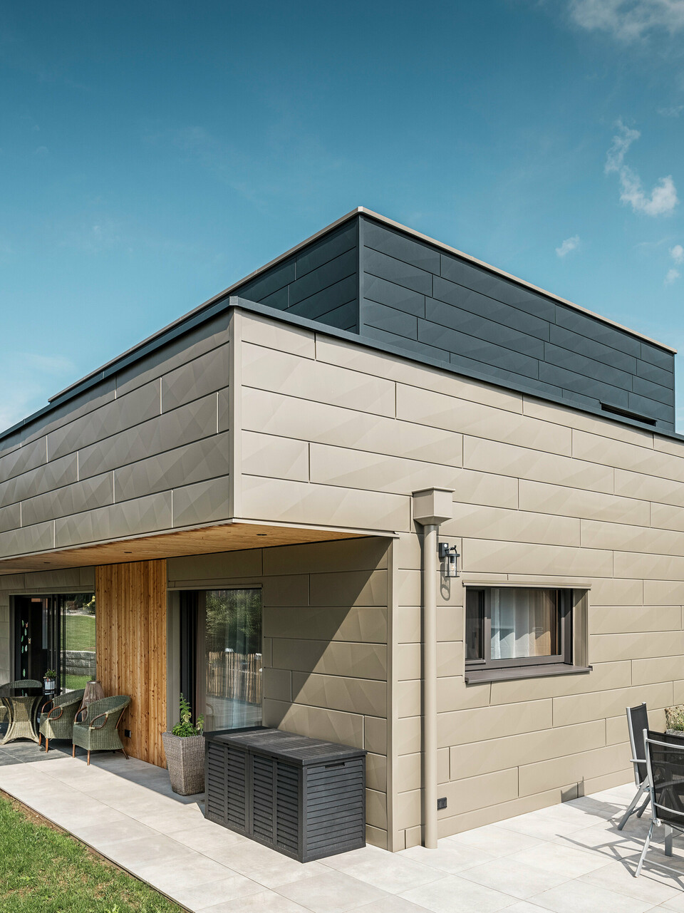 Ein modernes Einfamilienhaus in Wippenham, Österreich, mit einer zweifarbigen PREFA Aluminiumfassade, die aus den extravaganten Siding.X Fassadenpaneelen in Bronze und Anthrazit besteht. Die Fassade zeigt klar erkennbare, elegante Linien und Kantungen. Deutlich sichtbar ist die harmonisch integrierte PREFA Dachentwässerung mit Wasserfangkasten und Ablaufrohr, die sich nahtlos in das architektonische Gesamtkonzept einfügt. Im Vordergrund ist eine gemütliche Terrasse mit Gartenmöbeln zu sehen, die das moderne Design des neugebauten Hauses unterstreicht.