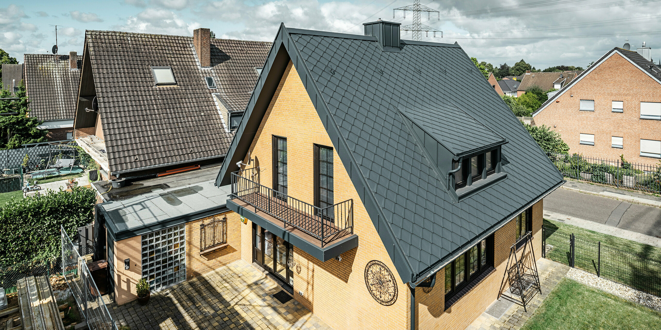 Einfamilienhaus in Tönisvorst mit einem spitzen Giebeldach, bedeckt mit PREFA Dachrauten 29×29 in P.10 Anthrazit. Das Dach weist eine klare Linienführung und Struktur auf. Die Dachrinne und das Ablaufrohr sind farblich abgestimmt und fügen sich nahtlos in das Design ein. Das Haus zeichnet sich durch gelbe Backsteinwände aus, die mit den dunklen Dachelementen kontrastieren, während ein Balkon und ein kleines Nebengebäude das äußere Erscheinungsbild vervollständigen.