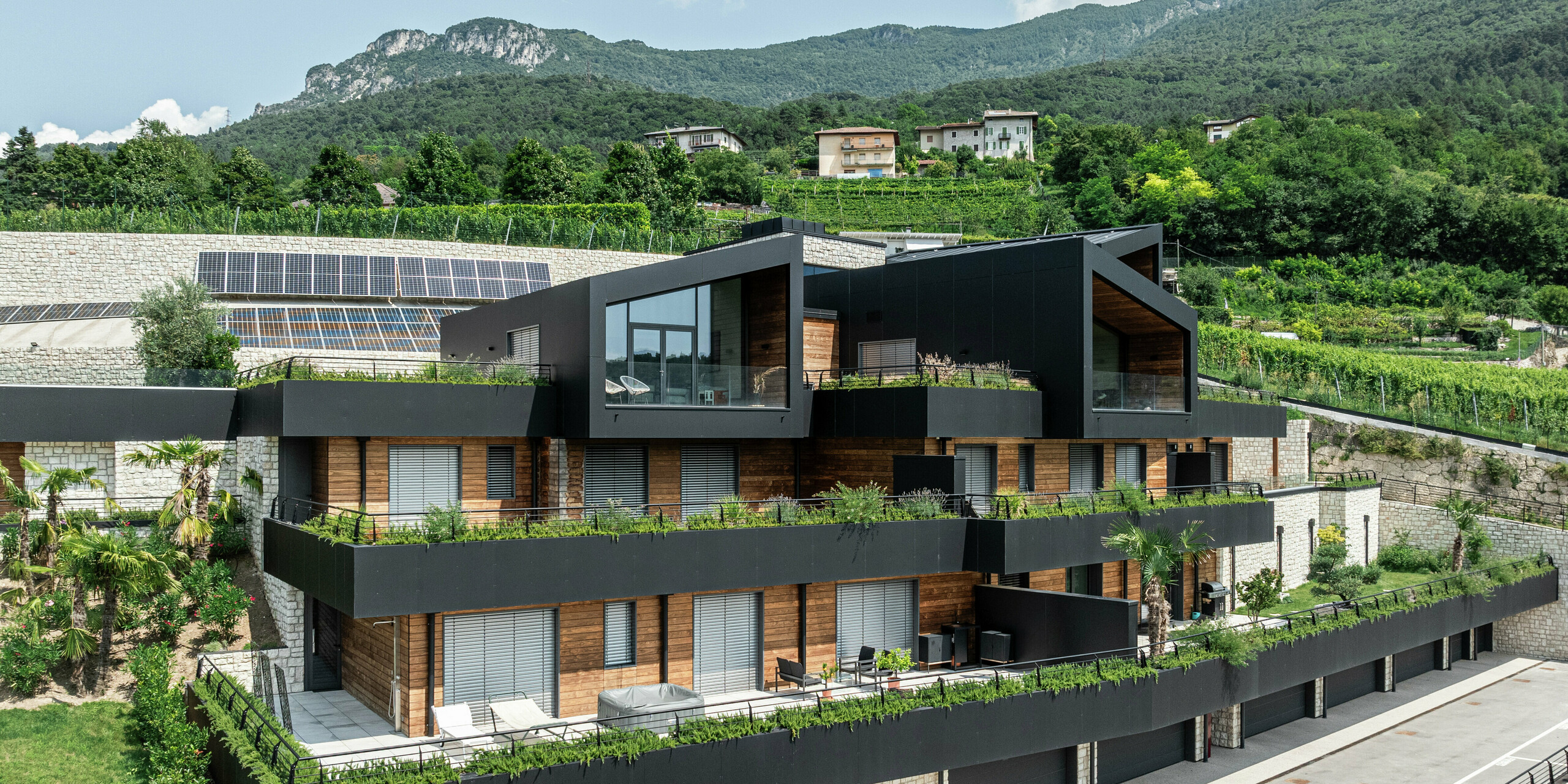 Moderne Wohnanlage in Hanglage mit nachhaltigem Design: Die Kombination aus Naturstein, Holz und der hochwertigen PREFA Aluminiumfassade in P.10 Schwarz verleiht dem Gebäude eine elegante und zeitlose Optik. Das PREFALZ Dach in P.10 Schwarz bietet optimalen Schutz und Langlebigkeit. Großzügige Terrassen, begrünte Dächer und Photovoltaikanlagen unterstreichen das nachhaltige Wohnkonzept. Die harmonische Einbettung in die Landschaft sowie die hochwertigen und pflegeleichten PREFA Materialien garantieren eine stilvolle und witterungsbeständige Architektur mit höchstem Wohnkomfort.
