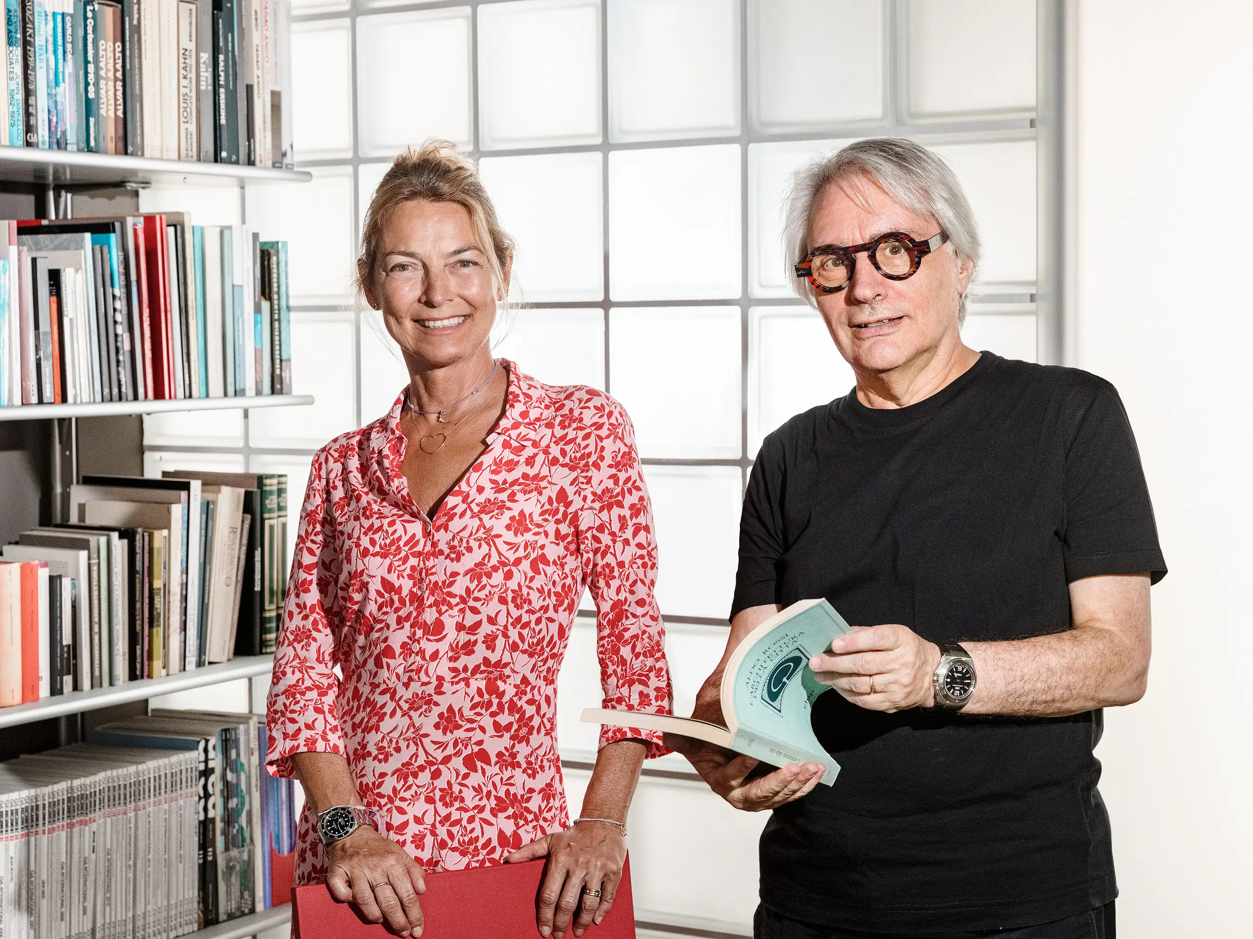 Der Bozener Architekt Claudio Lucchin und seine Büropartnerin Daniela Varnier posieren vor einer modernen Glaswand, die das Licht auf interessante Weise bricht. Claudio Lucchin ist ganz in Schwarz gekleidet und hält ein Buch in den Händen. Daniela Varnier trägt ein leuchtend rotes Oberteil und lächelt freundlich. Diese stilvolle Szene symbolisiert Innovation und Transparenz. Sie steht für futuristische Architektur – wie sie beispielsweise in der „Tempesti“-Kaserne in Corvara in Südtirol realisiert wurde.