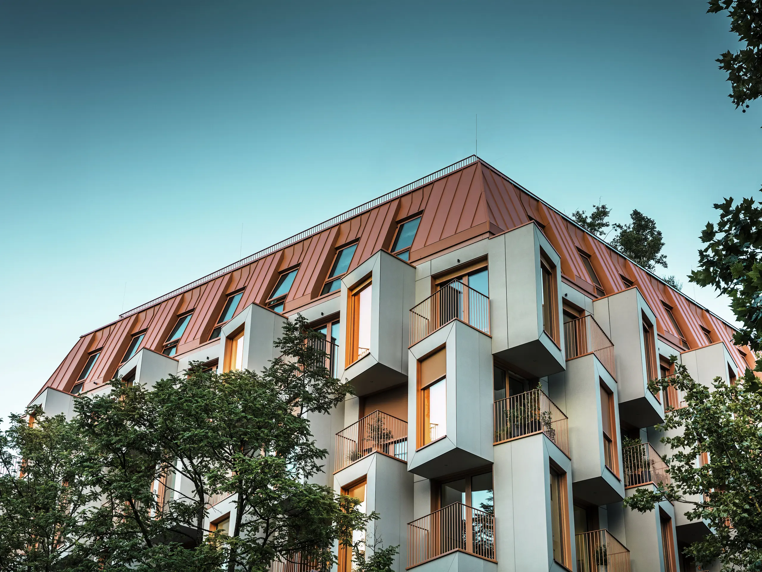 Architektonisch markante Fassade des Wohnprojekts Van B in München mit auffälligen Erkerfenstern und einem PREFALZ-Aluminiumdach in der Sonderfarbe IGP Orange HDP. In der Abenddämmerung erstrahlen die Fenster im Licht. Die moderne Dachlösung vereint höchste Witterungsbeständigkeit mit elegantem Erscheinungsbild und setzt farblich einen Akzent im Stadtbild. Das Zusammenspiel von glasfaserverstärktem Beton, kupferroten Fensterrahmen und Aluminiumdach schafft eine dynamische, langlebige und nachhaltige Gebäudehülle – ideal für zukunftsorientiertes Bauen.