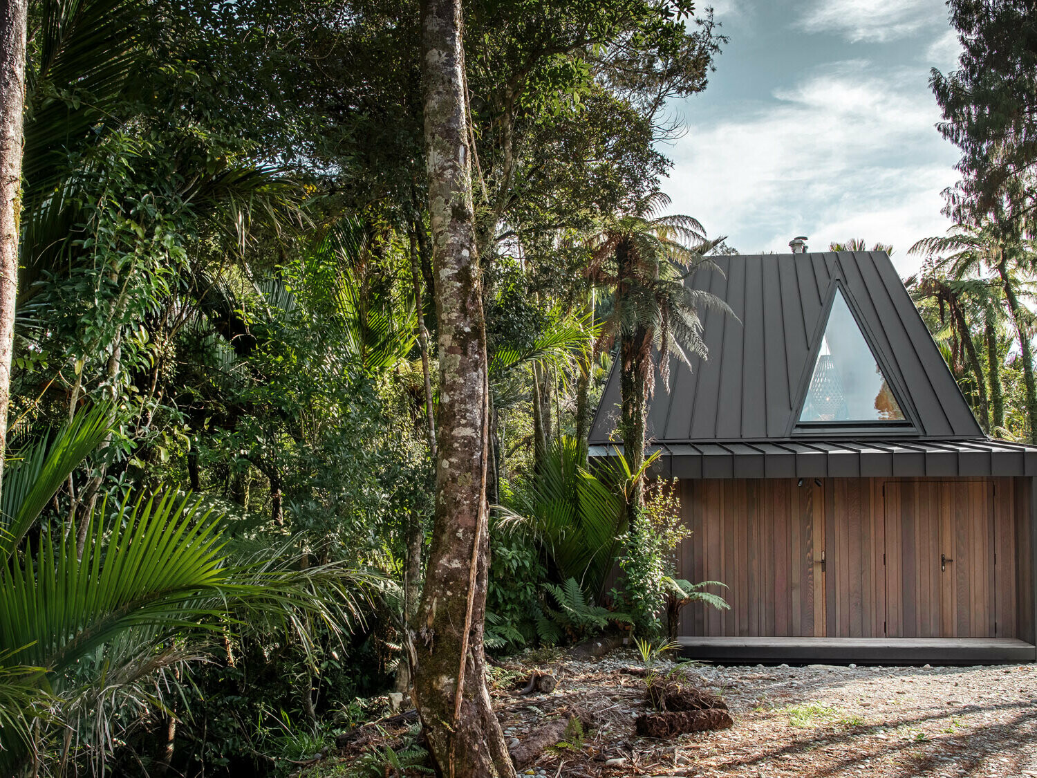 Seitlicher Blick auf das von Fabric realisierte Biwak mit dem ausgefallenen, schwarzgrauen Prefalz Dach, das sich zwischen Nikau-Palmen im Punakaiki Urwald versteckt.