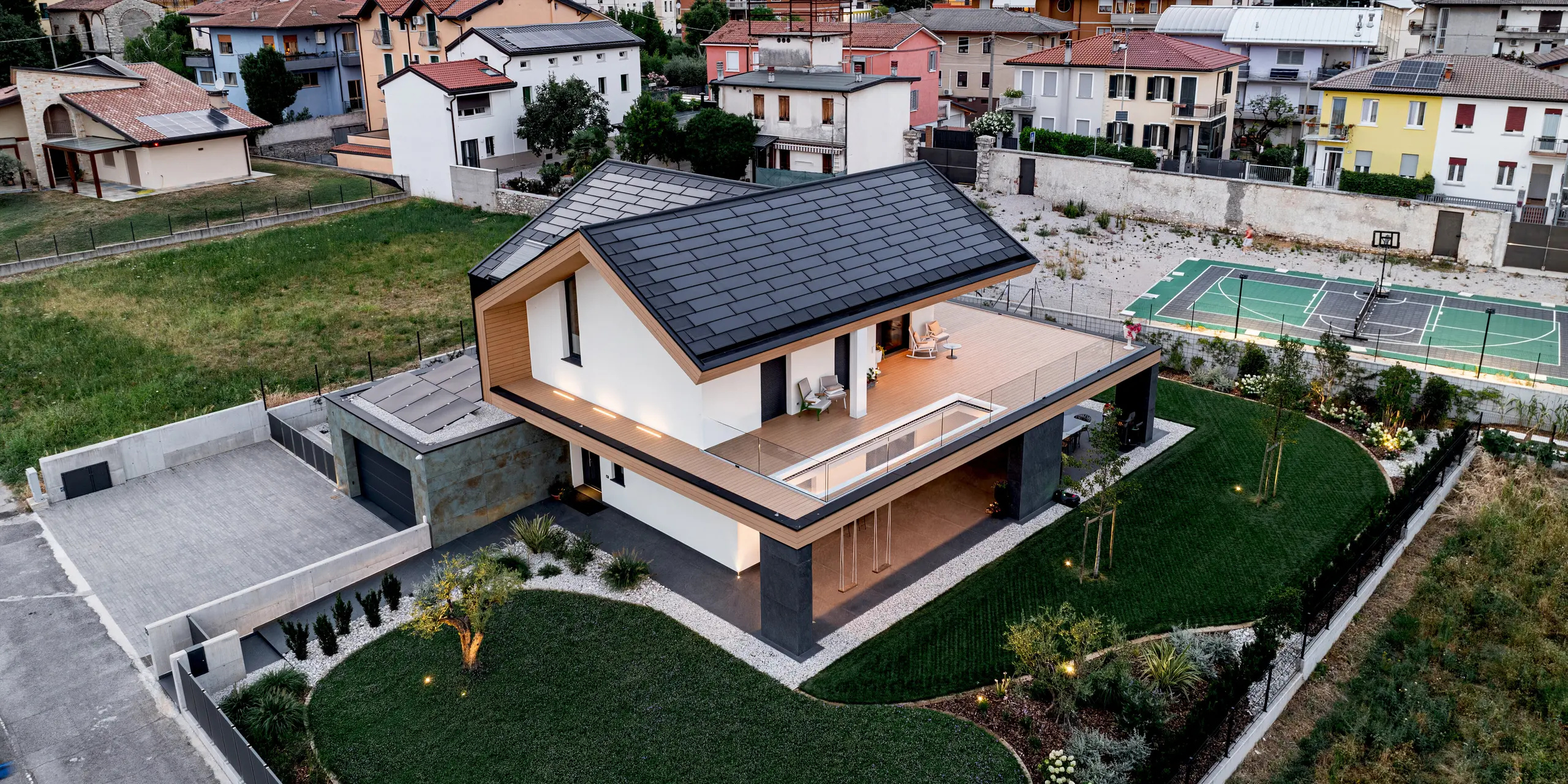 Luftaufnahme eines modernen Einfamilienhauses in Vicenza mit PREFA Dachplatte R.16 und PREFA Solardachplatte in P.10 Schwarz. Die markante Dachgestaltung hebt sich harmonisch von der grünen Berglandschaft ab. Die langlebigen Aluminiumprodukte verbinden elegantes Design, nachhaltige Energiegewinnung und zuverlässigen Schutz für eine zukunftsorientierte Architektur.