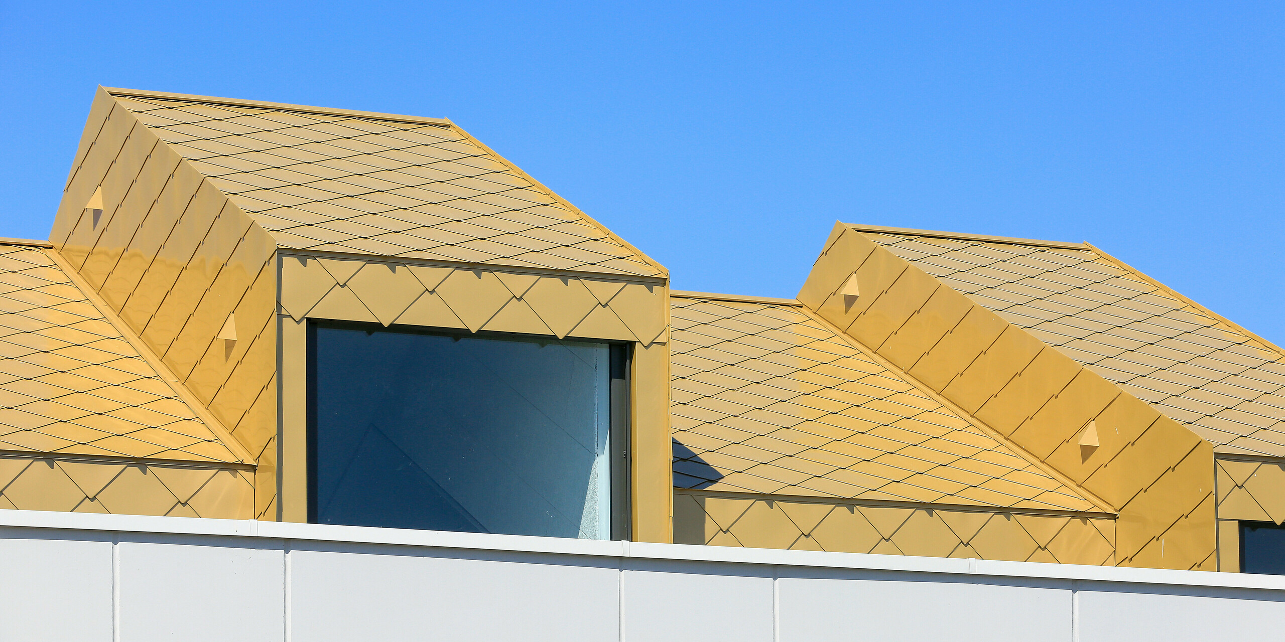 Nahaufnahme eines Dachfensters des Gemeinde- und Kulturzentrums Fessenheim in Frankreich. Das Fenster ist umhüllt von PREFA Dach- und Wandrauten 44 × 44 in Sparkling Gold.