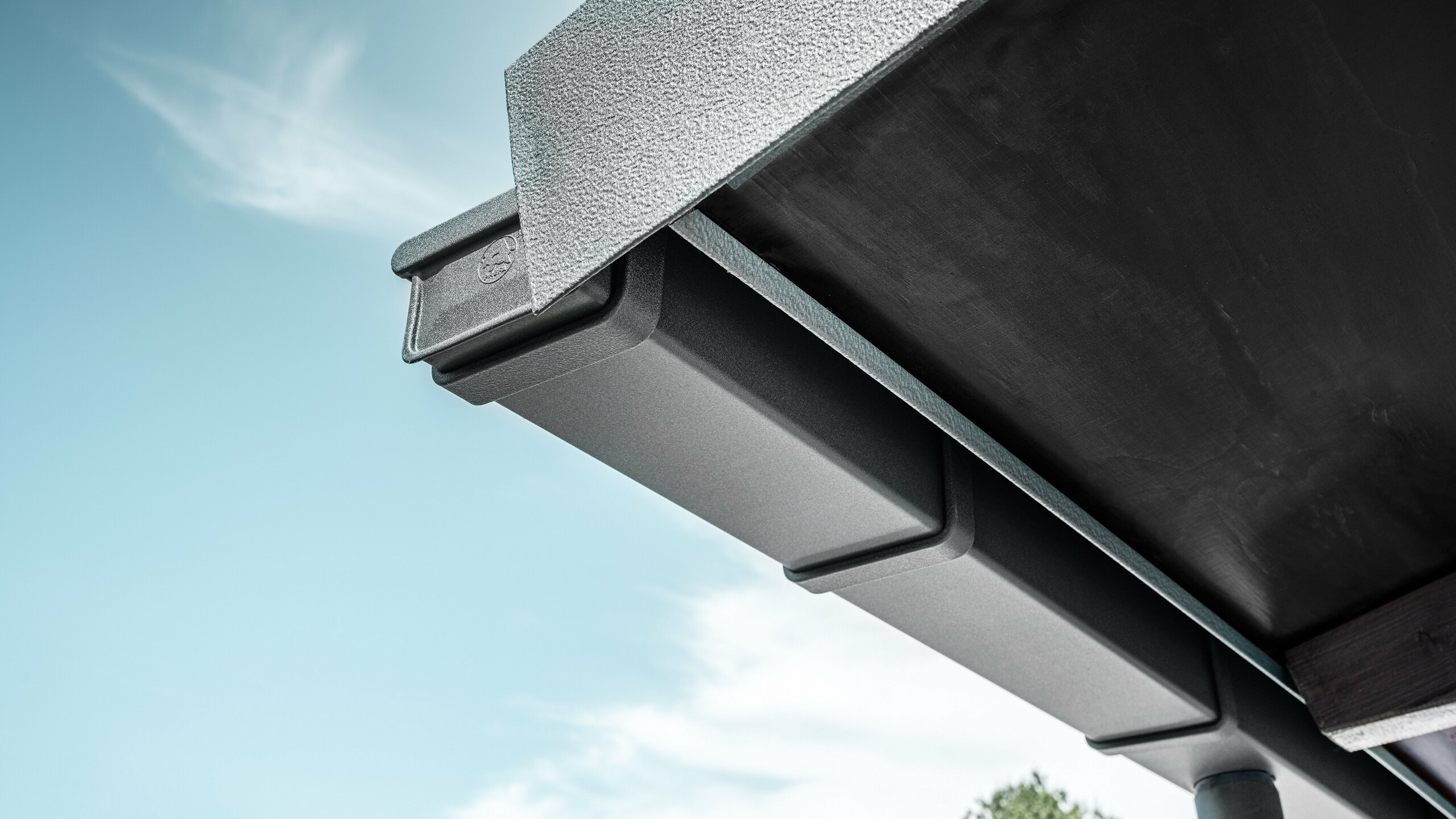 Detailaufnahme einer eckigen PREFA Kastenrinne in Anthrazit aus Aluminium mit strukturiertem Abschlussblech, montiert an der Dachkante eines Gebäudes vor blauem Himmel mit leichter Bewölkung.