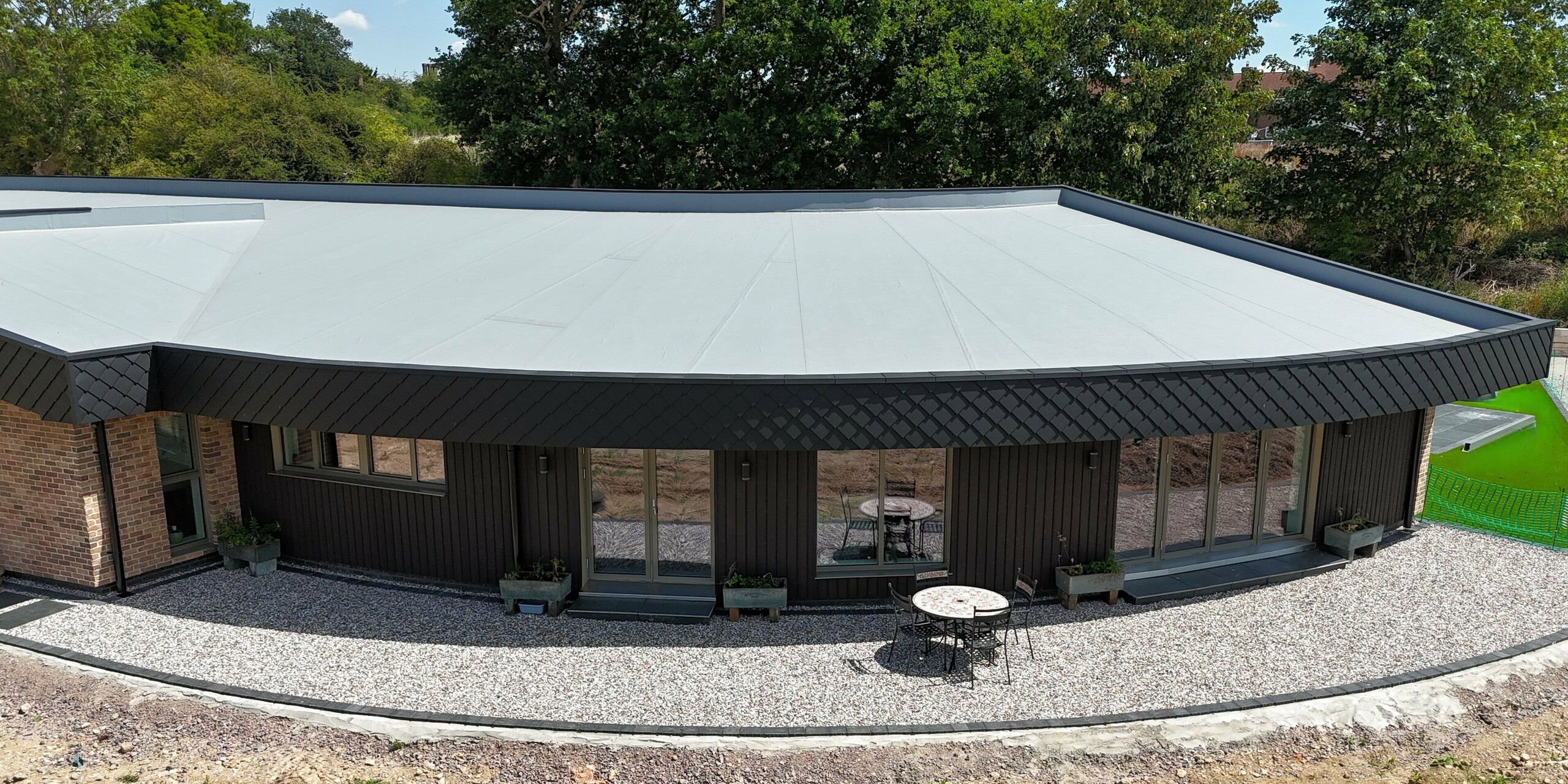 Modernes Einfamilienhaus mit markanter Gebäuderundung und stilvoller Fassadengestaltung. Die Kombination aus dunklen Holzpaneelen und der kleinformatigen PREFA Wandraute 20 × 20 in P.10 Anthrazit verleiht der Fassade einen eleganten, dynamischen Look. Die Wandrauten aus Aluminium sind nicht nur ein optisches Highlight, sondern bieten auch Vorteile wie Langlebigkeit, Witterungsbeständigkeit und Pflegeleichtigkeit. Große Glasflächen öffnen den Wohnraum zur Natur, während die klare Linienführung und die hochwertige Materialwahl nachhaltige Architektur perfekt in Szene setzen.