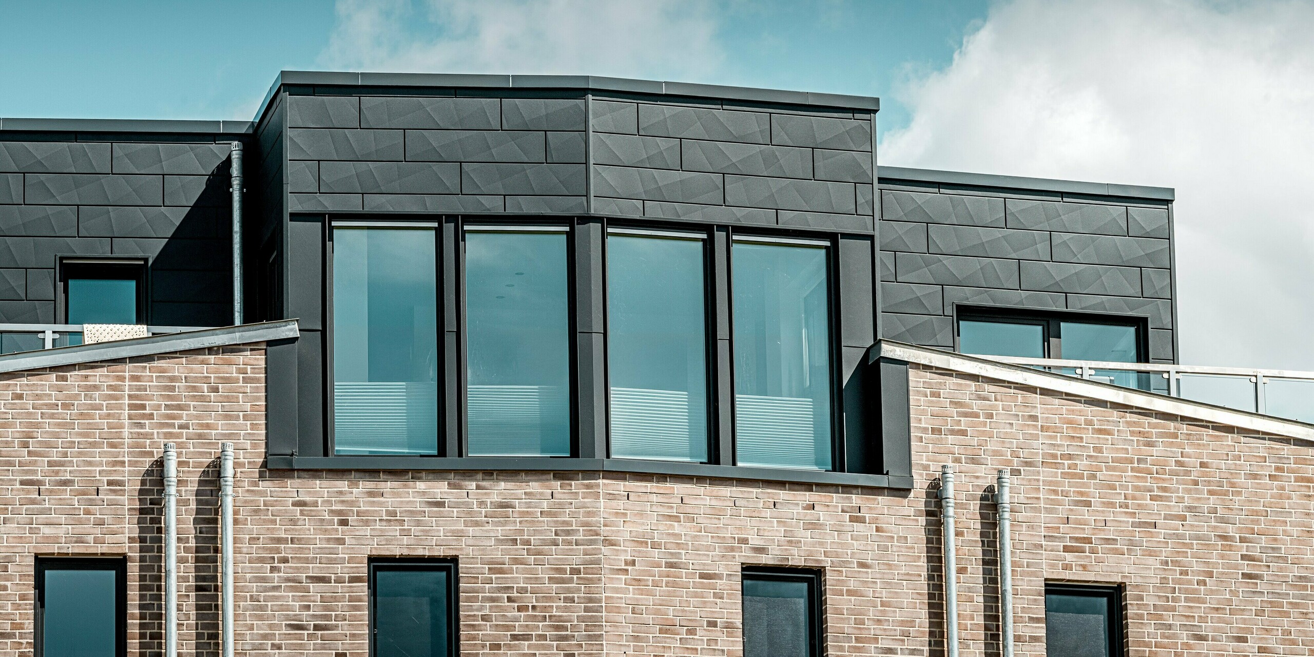 Auf dem Detailfoto ist das markante Dachgeschoss der Wohnanlage „Hafen zur Freiheit“ zu sehen, bei dem die PREFA Siding.X-Fassadenpaneele in P.10 Anthrazit besonders hervorstechen. Die reliefartige Aluminiumstruktur verleiht dem oberen Gebäudeteil eine kraftvolle, moderne Ausstrahlung und bildet einen spannenden Kontrast zur hellen Klinkerfassade darunter. Die großzügigen Fensterflächen spiegeln den Himmel wider und betonen die hochwertige Architektur. Dank der robusten und korrosionsbeständigen PREFA Aluminiumpaneele bleibt die Fassade selbst in der rauen Küstenlage langlebig und optisch ansprechend.