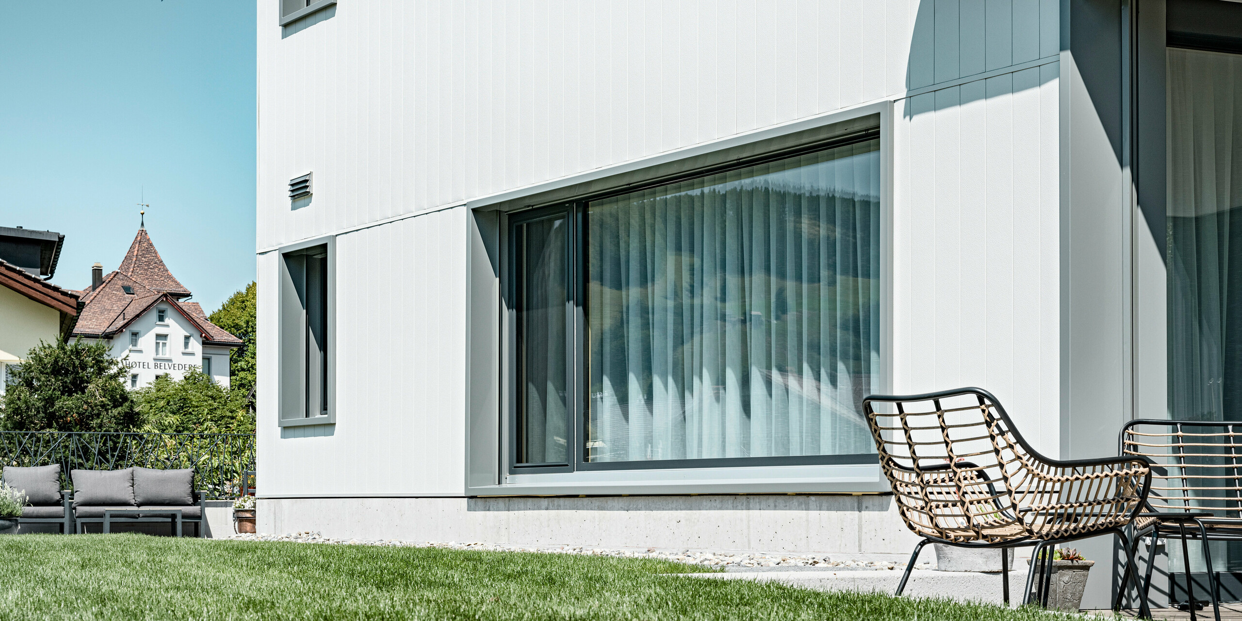 Harmonische Verbindung von Funktionalität und Design: Das Mehrfamilienhaus in Weissbad, Schweiz, zeigt seine klare, moderne Linienführung durch die PREFA Aluminiumfassade in P.10 Prefaweiß. Die vertikal montierten PREFA Sidings unterstreichen das schlichte, aber stilvolle Erscheinungsbild des Gebäudes, während die großen Fensterfronten reichlich Tageslicht in die Wohnräume lassen. Die Fassade bildet einen ruhigen Hintergrund für die entspannende Gartensitzecke.