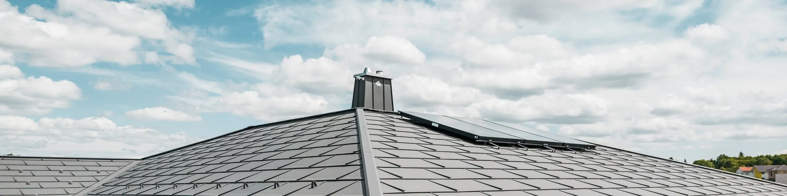 Detailansicht eines PREFA Aluminiumdachs auf einem Einfamilienhaus in Ilmenau, Thürigen. Das Dach wurde überwiegend mit PREFA Dachplatten R.16 in P.10 Dunkelgrau eingedeckt. Die kleinformatigen Aluminiumdachplatten wurden fachmännisch verlegt und verleihen dem Bungalow einen modernen Look. Das Bild zeigt die Präzision des PREFA Dachsystems, inklusive der Schneefangsysteme, die für zusätzliche Sicherheit sorgen. Die harmonische Integration der Hängerinnen und Ablaufrohre des PREFA Dachentwässerungssystems betont die ästhetische und funktionale Qualität des Gebäudes. Am linken Bildrand sind Stehfalzstreifen zu sehen, welche mit dunkelgrauem PREFALZ auf der direkt anschließenden Garage montiert wurden.