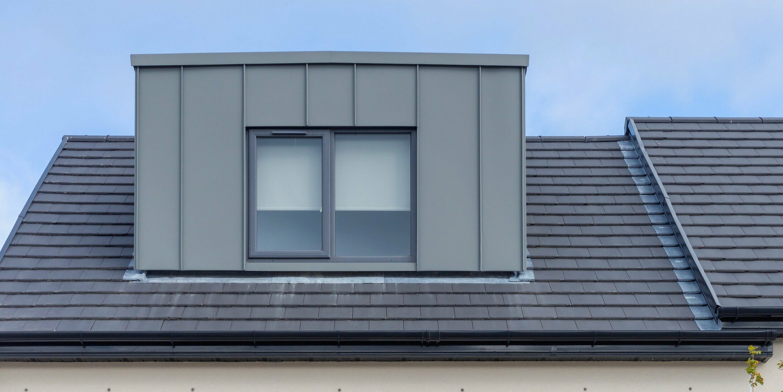 Das Bild zeigt ein modernes Reihenhaus in Churchfields, dessen Gaube mit hochwertigem PREFALZ in P.10 Hellgrau verkleidet ist. Die klare Stehfalzstruktur der Aluminiumoberfläche setzt einen markanten Akzent über dem dunklen Ziegeldach und verleiht dem Gebäude eine elegante, zeitgemäße Ausstrahlung. Die langlebige, korrosionsbeständige PREFA Verkleidung sorgt für ein harmonisches Gesamtbild und reduziert den Wartungsaufwand erheblich – ein großer Vorteil für Wohnanlagen dieser Größe.