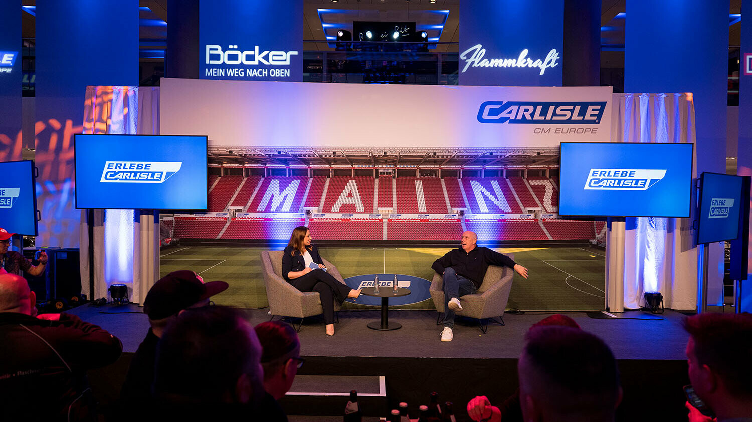 Moderierte Gesprächsrunde auf der Bühne des Carlisle Branchenevents in der Mainz Arena. Zwei Gesprächspartner diskutieren vor Publikum, im Hintergrund ist eine Stadionkulisse mit der Aufschrift ‚MAINZ‘ sowie das Branding von Carlisle CM Europe und ‚Erlebe Carlisle‘ zu sehen.