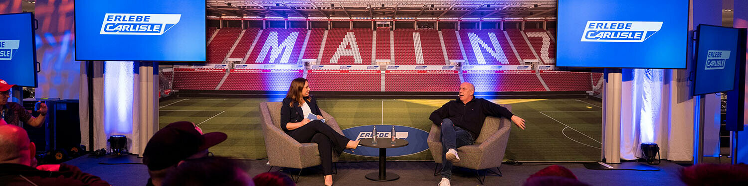 Moderierte Gesprächsrunde auf der Bühne des Carlisle Branchenevents in der Mainz Arena. Zwei Gesprächspartner diskutieren vor Publikum, im Hintergrund ist eine Stadionkulisse mit der Aufschrift ‚MAINZ‘ sowie das Branding von Carlisle CM Europe und ‚Erlebe Carlisle‘ zu sehen.