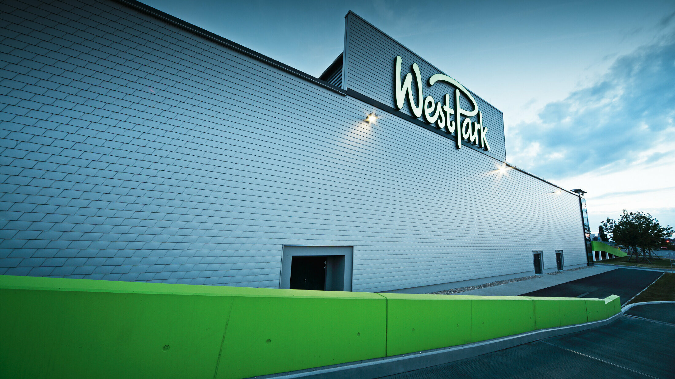Moderne Fassade eines Einkaufszentrums mit großem Schriftzug „WestPark“. Die Außenwand ist mit silbermetallicfarbenen Aluminium-Schindeln verkleidet und wirkt durch die gleichmäßige Struktur elegant und technisch. Im Vordergrund verläuft eine leuchtend grüne Mauer entlang des Gehwegs. Das Gebäude ist in der Abenddämmerung beleuchtet.