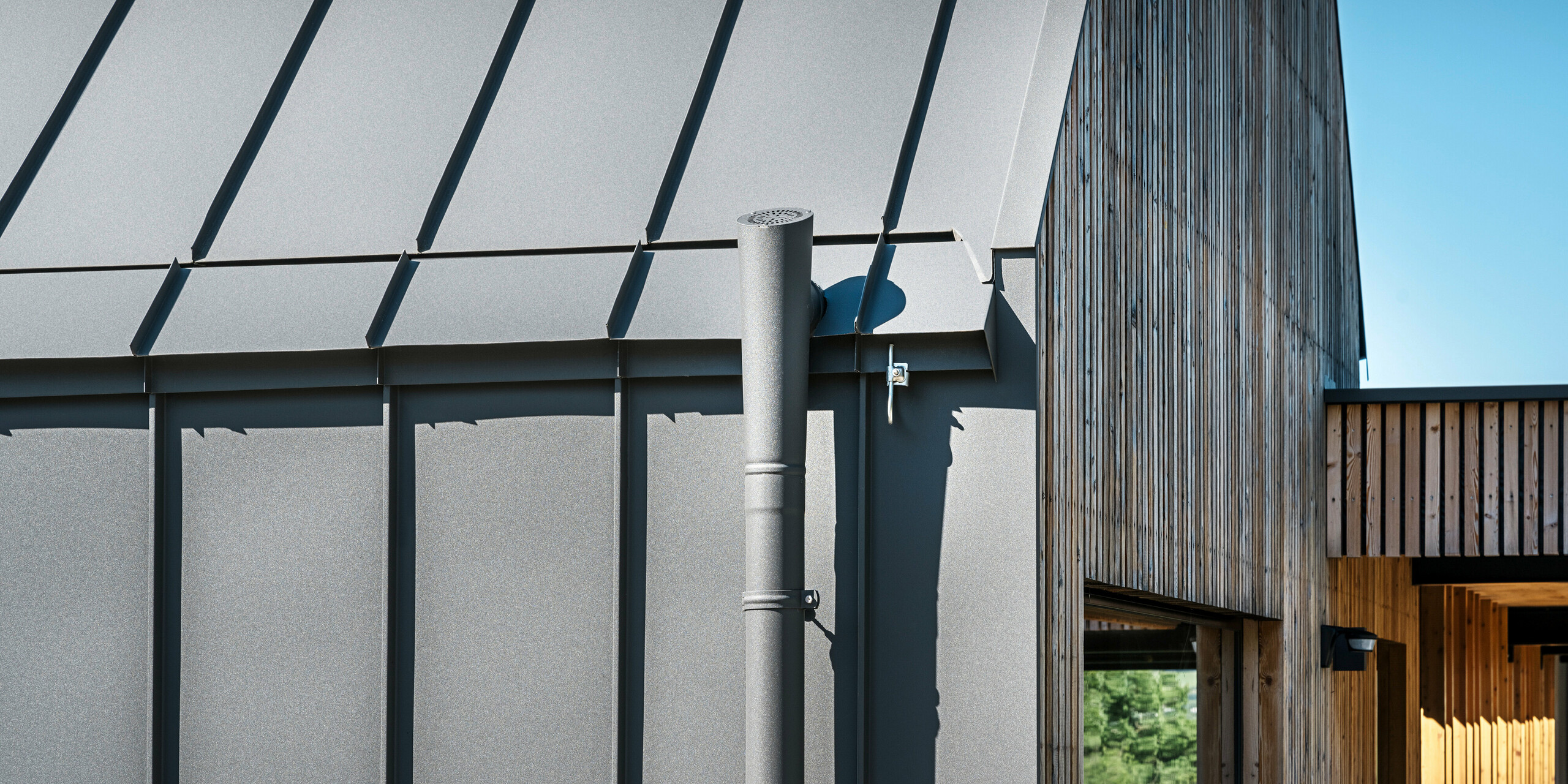 Detailní záběr návrhu střechy a fasády rodinného domu v rakouském Pogier s vysoce kvalitním PREFALZ v P.10 tmavě šedá. Odvodnění střechy je realizováno pomocí barevně sladěného odtokového potrubí, které se plynule integruje do elegantního celkového vzhledu. Kombinace precizních linií střechy či fasády a přírodního dřevěného obkladu vstupního prostoru v pozadí dokresluje estetickou souhru moderních a tradičních stavebních materiálů.