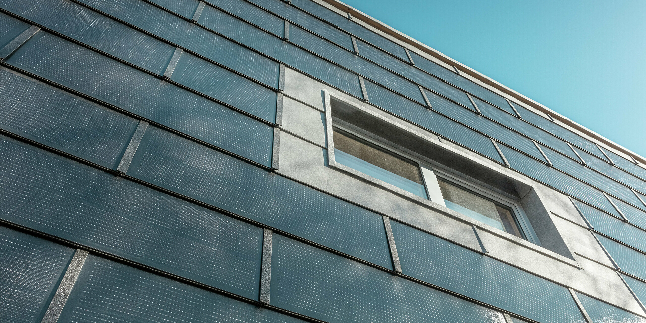 Die schräg aufgenommene Fassade der Spenglerei RÜMA in Vorarlberg ist mit PREFA Solardachplatten verkleidet. Die grauen Aluminiumelemente und die integrierten Photovoltaikmodule umrahmen ein Fenster. Im Hintergrund ist ein strahlend blauer Himmel zu erkennen.