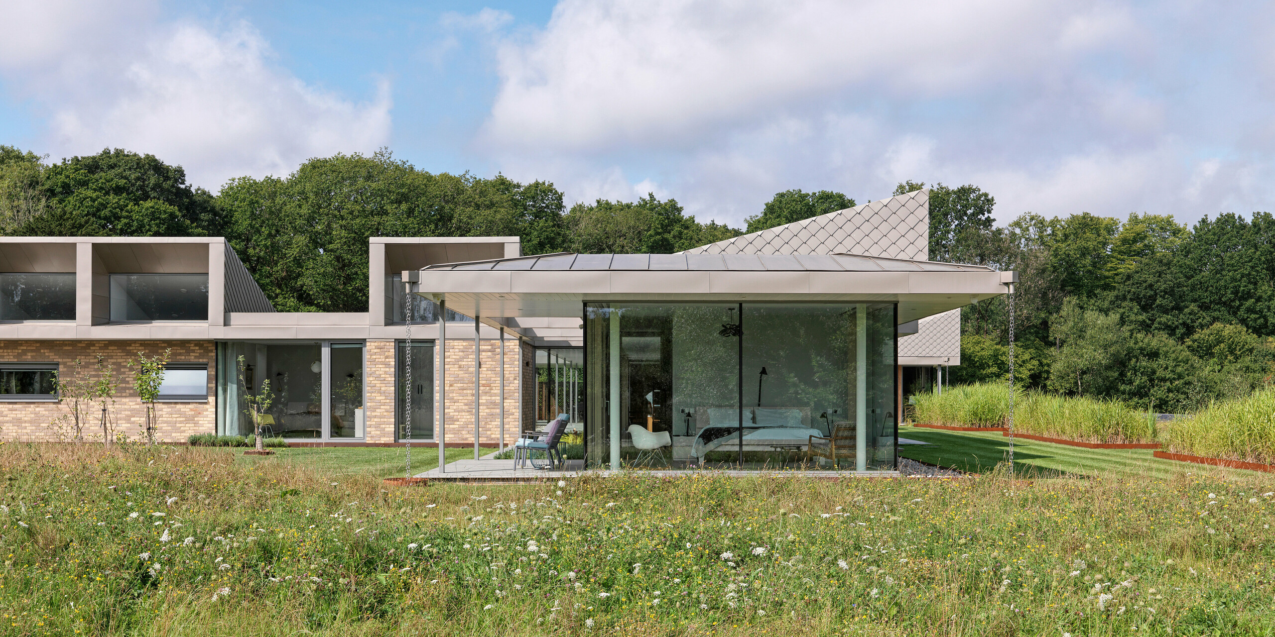 Seitenansicht des modernen Bungalows in West Sussex mit PREFALZ Aluminiumdach und akzentuierten Fassadenbereichen aus PREFA Wandrauten 20 × 20 in P.10 Bronze. Die raumhohen Glasfronten der Wohnbereiche öffnen sich zur Wildblumenwiese im Vordergrund und schaffen eine nahtlose Verbindung zwischen Innenraum und Natur. Das langlebige PREFA Dach- und Fassadensystem verleiht dem Gebäude nicht nur Schutz, sondern auch architektonische Eleganz.