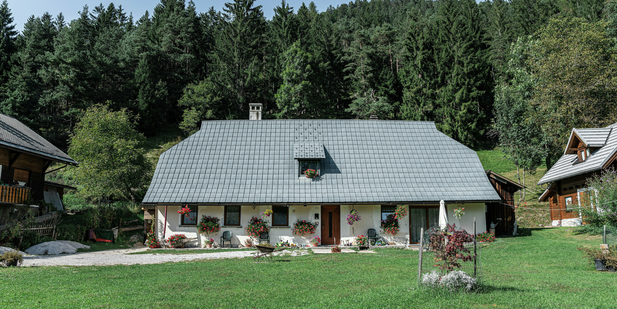 Ein malerisches Einfamilienhaus in Tarvis, Italien, seit 1972 mit robusten PREFA Dachplatten eingedeckt: Das große Krüppelwalmdach mit der Aluminiumeindeckung schützt das Gebäude seit Jahrzehnten zuverlässig vor den alpinen Witterungseinflüssen. Die Fassade zieren bunte Blumenkästen, der gepflegte Garten mit Sitzecke und kleiner Feuerstelle lädt zum Verweilen ein. Umgeben von dichten Nadelbäumen und traditionellen Holzhäusern fügt sich das Haus harmonisch in die alpine Naturlandschaft ein.
