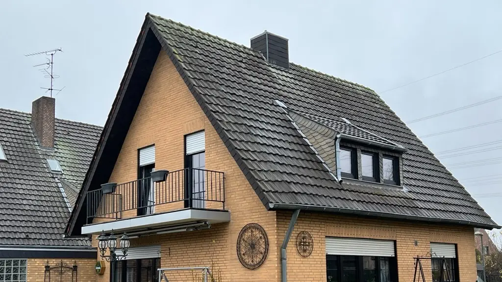 Einfamilienhaus aus gelbem Klinker mit steilem Satteldach und dunkler Dacheindeckung vor der Dachsanierung; auf der Gartenseite mit Balkon, Dachgaube, Terrasse mit Sitzmöbeln und Rasenfläche im Vordergrund.