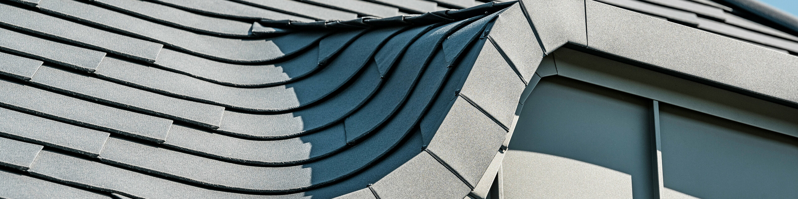 Nahaufnahme einer geschwungenen Hechtgaube, gedeckt mit PREFA Dachschindeln in P.10 Anthrazit, die sich durch ihre Flexibilität bei der Dachgestaltung auszeichnen. Die Detailansicht zeigt die präzise Verlegung und die hohe Anpassungsfähigkeit der Aluminiumschindeln an komplexe Dachformen, was die handwerkliche Qualität und die innovative Technologie von PREFA unterstreicht.