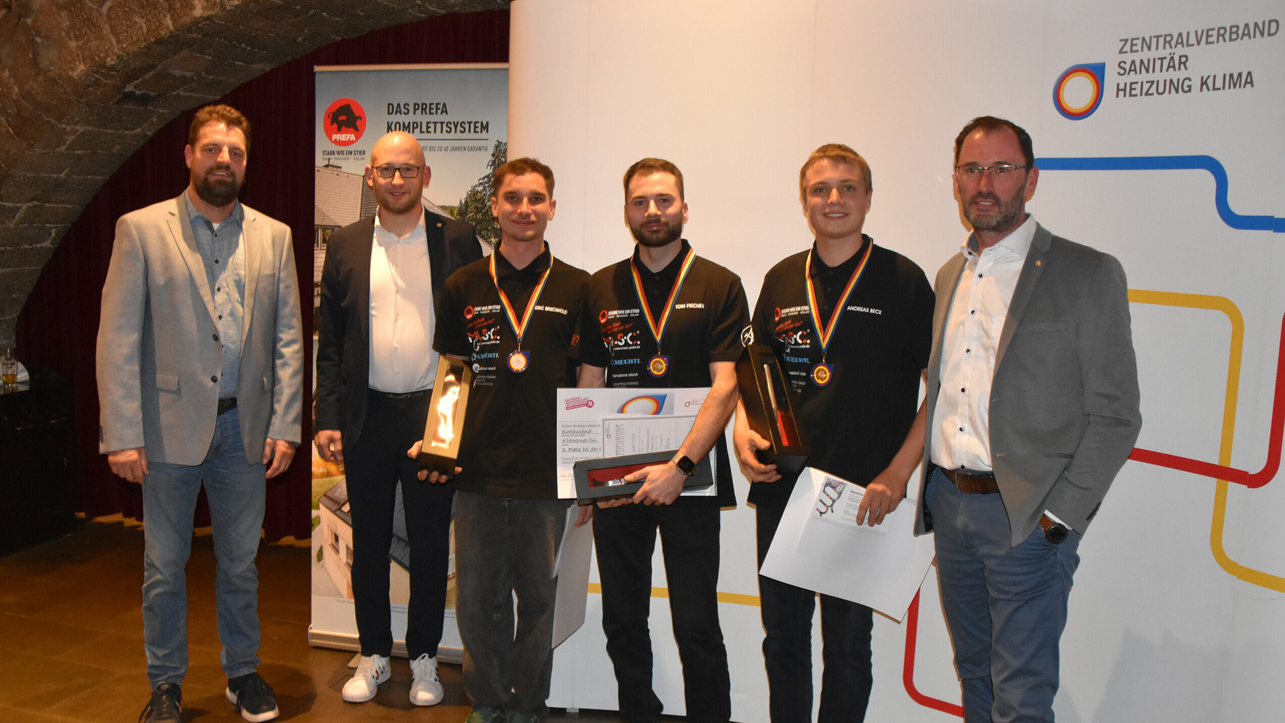 Die drei Erstplatzierten zeigen ihre Medaillen und Pokale nach dem Wettbewerb. Sie stehen mit den Vertretern des ZVSHK und PREFA, die das Ereignis begleiteten.