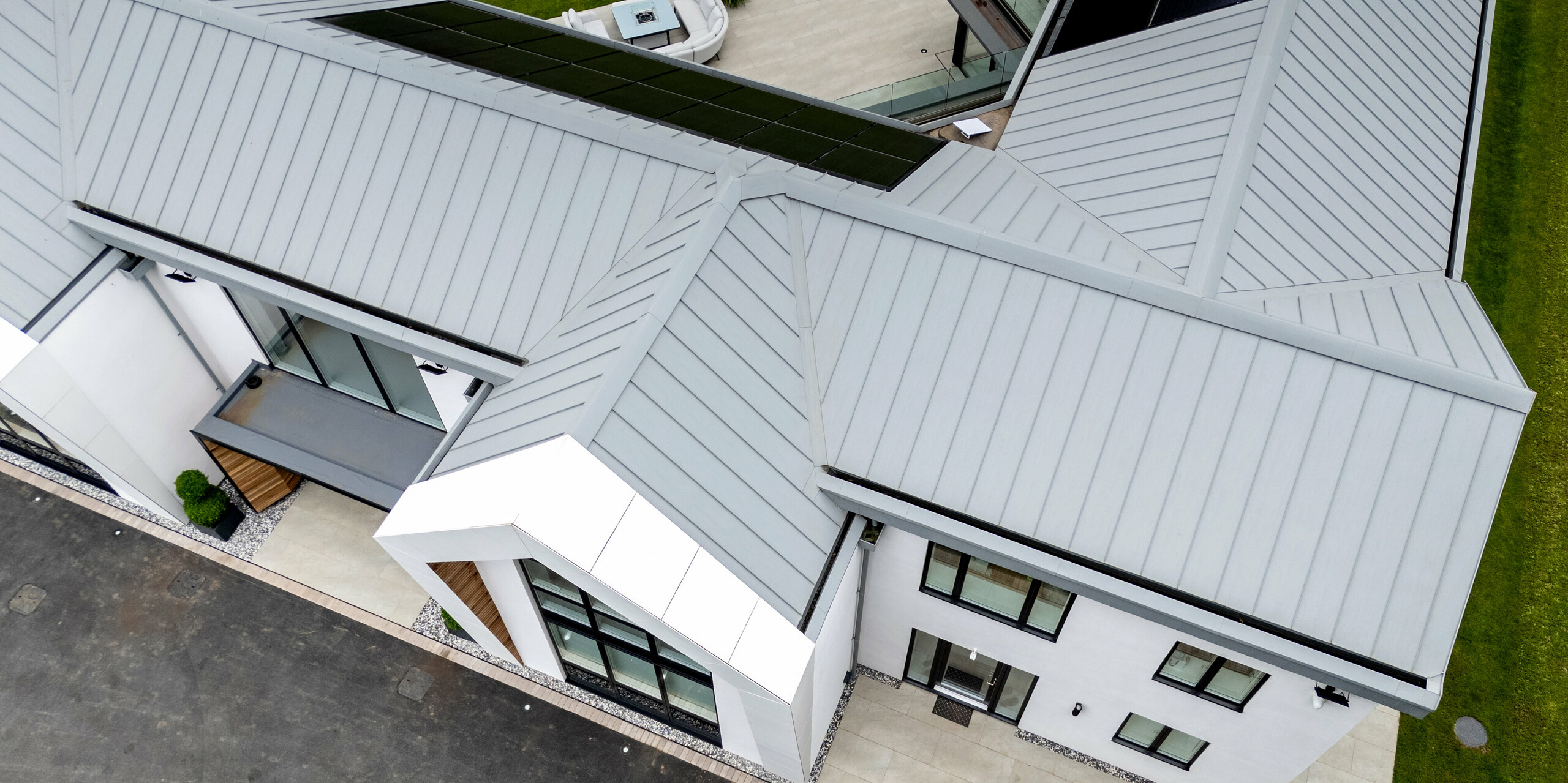 Die Ansicht aus der Vogelperspektive zeigt das elegante PREFALZ-Dach in der Farbe Patinagrau der modernen Villa in Draycott in the Clay. Die Stehfalzstruktur verleiht dem Blechdach ein klares, modernes Aussehen und fügt sich perfekt in die Umgebung ein. Das mit Photovoltaik-Modulen ausgestattete Dach unterstreicht die umweltfreundliche Ausrichtung des Hauses. Die symmetrische Anordnung und die präzise Ausführung des Daches sorgen für ein ästhetisches Gesamtbild, das Funktionalität und Design vereint.