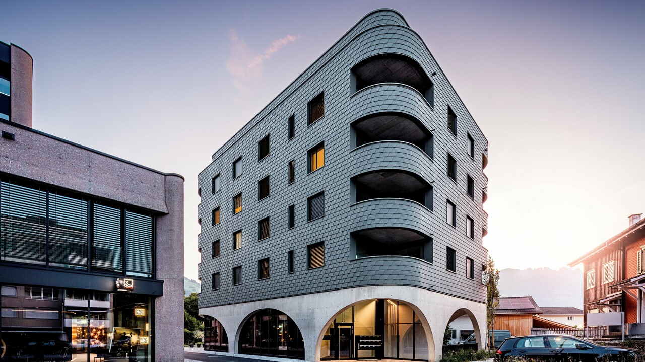 Modernes Mixed-Use-Gebäude in Buchs mit markant gerundeter, dreieckiger Form und durchgehender Fassade aus PREFA Wandschindeln in P.10 Hellgrau. Die schuppenartige Aluminiumhaut umhüllt das Gebäude wie eine Reptilienstruktur und betont die fließenden Rundungen sowie die tief eingeschnittenen Balkone. Über dem massiven Sockel mit eleganten Betonarkaden wirkt die leichte, witterungsbeständige PREFA Fassade besonders ausdrucksstark. Präzise verlegte Aluminiumschindeln sorgen für eine homogene, langlebige und wartungsarme Gebäudehülle mit starkem Wiedererkennungswert.