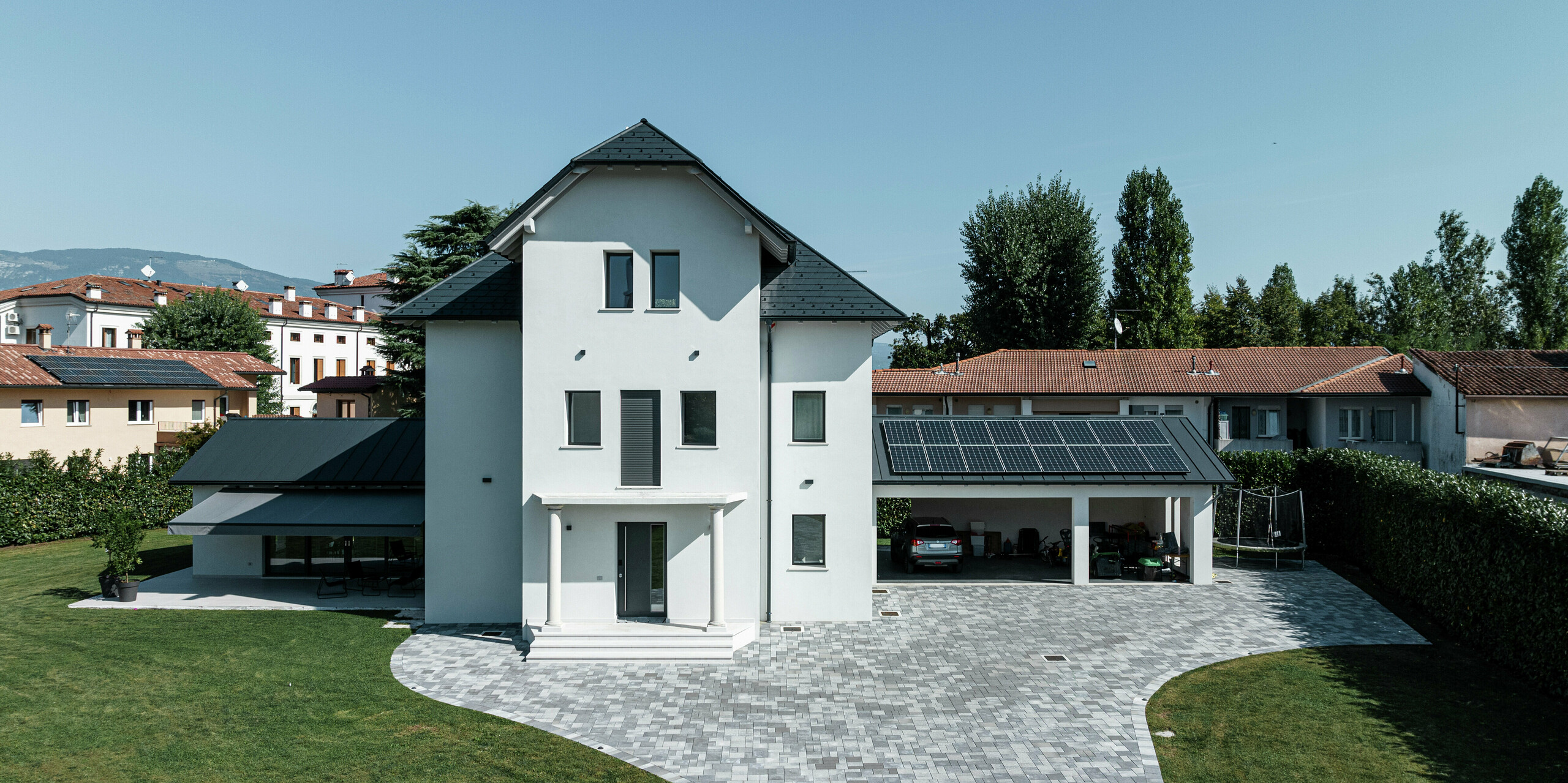 Frontansicht eines modernen Einfamilienhauses in Marano Vicentino, Italien, mit widerstandsfähigen PREFA Dachschindeln P.10 Anthrazit. Die angrenzenden Nebengebäude links und rechts sind mit bewährtem Doppelstehfalz aus Aluminium eingedeckt - ebenfalls in Anthrazit. Über der Garage sorgt eine Photovoltaikanlage für Strom aus erneuerbarer Energie. Die helle Putzfassade und das dunkle Dach bilden einen eleganten Kontrast an dem dreigeschossigen Einfamilienhaus.