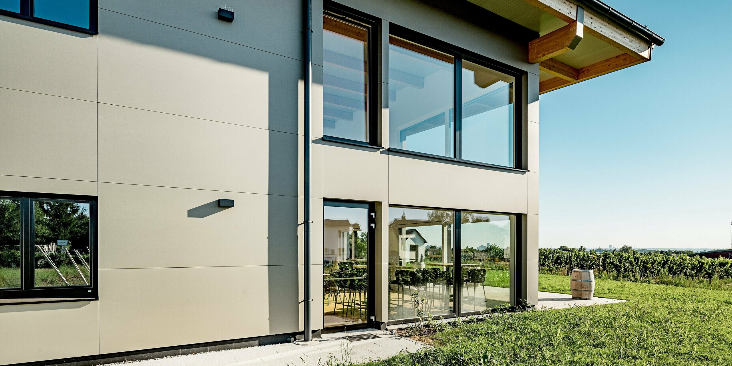 Seitlicher Fassadenausschnitt des Weinguts Nigl in Perchtoldsdorf mit großzügigen Fensterflächen und PREFA Aluminium Verbundplatten in P.10 Prefabronze. Die klare Linienführung der Fassadenelemente verleiht dem Gebäude ein modernes und aufgeräumtes Erscheinungsbild. Sichtbar ist auch das elegante Zusammenspiel aus natürlichen Materialien und der widerstandsfähigen Aluminiumfassade. Die bodentiefen Fenster bringen viel Licht ins Innere und eröffnen den Blick auf die angrenzenden Weingärten. Die langlebigen, witterungsbeständigen PREFA Platten bieten optimalen Schutz und unterstreichen gleichzeitig die stilvolle Architektur des modernen Gewerbebaus.