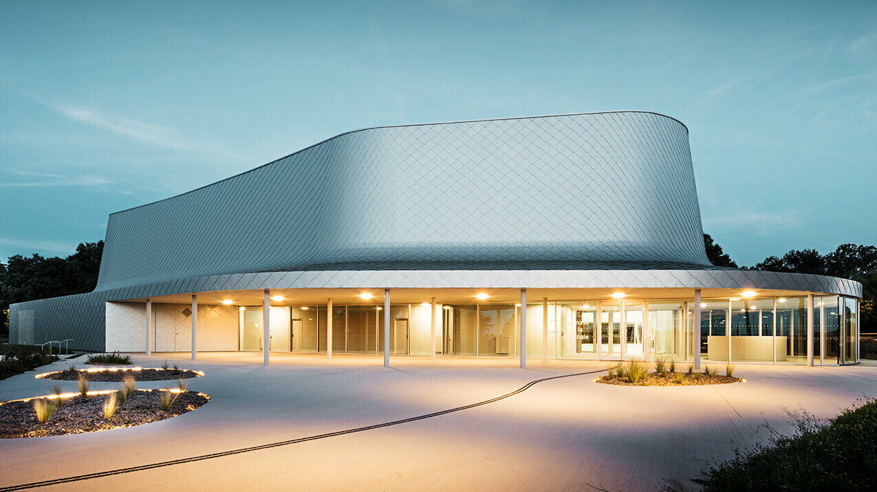 Seitliche Abendaufnahme der von Bohuon Bertic Architectes entworfenen Sporthalle in Basse-Goulaine mit Beleuchtung.