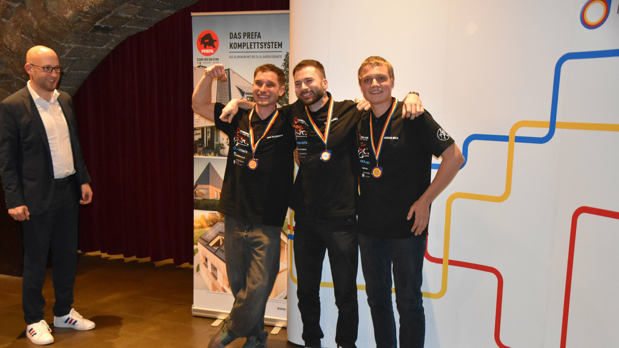 Die besten drei Teilnehmer des Wettbewerbs, alle mit Medaillen, feiern ihren Erfolg. Im Hintergrund ist ein Vertreter des ZVSHK zu sehen, der die Auszeichnungen übergibt.