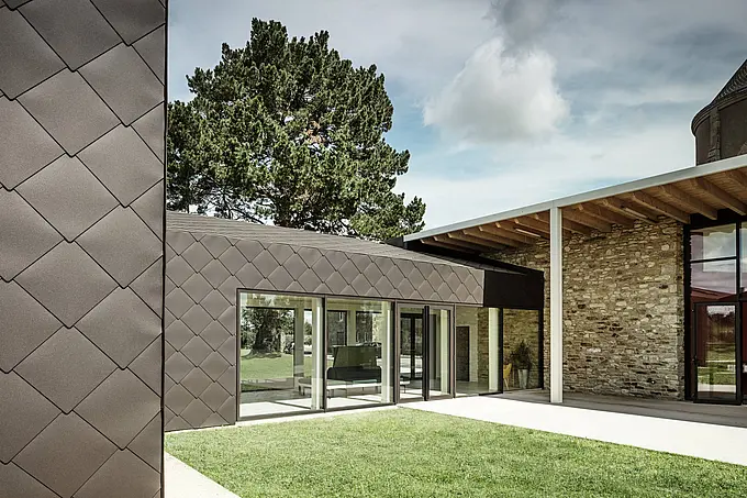 Das Bild zeigt eine moderne Gebäudefassade, die mit PREFA-Dach- und Wandelementen (44 × 44 mm) in Walnussbraun verkleidet ist. Die Rautenstruktur der Aluminiumverkleidung verleiht der Fassade ein elegantes und dynamisches Erscheinungsbild. Große Glasflächen öffnen den Innenraum zur umgebenden Natur, während die Kombination aus Steinmauern und moderner Verkleidung einen harmonischen Übergang zwischen traditioneller und zeitgenössischer Architektur schafft. Ein grüner Rasen unterstreicht zusätzlich den natürlichen und einladenden Charakter des Gebäudes.