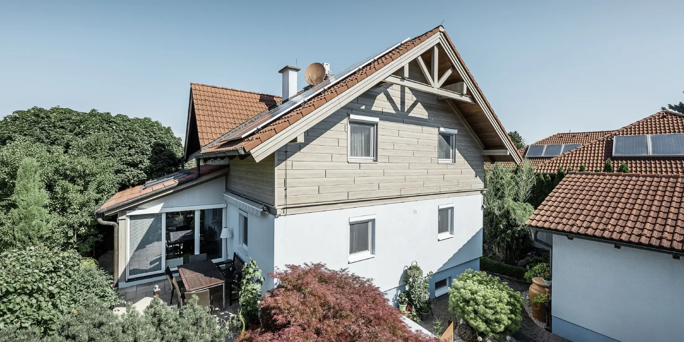 Einfamilienhaus mit Satteldach und roter Dacheindeckung, kombiniert mit einer modernen Fassadengestaltung. Der obere Giebel ist mit beige-grauen Aluminium-Fassadenpaneelen von PREFA verkleidet, während das Erdgeschoss eine weiße Putzfassade hat. Auf dem Dach befinden sich Solarmodule. Das Haus liegt in einem grünen Garten mit Terrasse, Gartenmöbeln und üppiger Bepflanzung, umgeben von weiteren Wohnhäusern mit ähnlicher Dacharchitektur.