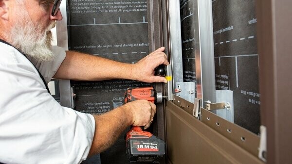 Das Bild zeigt eine Person, die einen Akkuschrauber benutzt, um den Sturmsicherungsclip an der Fassade zu befestigen. Man sieht auch einen Kugelkopf, der als Montagehilfe dient. Es ist eine Szene aus einer professionellen Handwerksarbeit, mit Fokus auf Präzision und sachgerechter Ausführung.