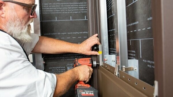 Das Bild zeigt eine Person, die einen Akkuschrauber benutzt, um den Sturmsicherungsclip an der Fassade zu befestigen. Man sieht auch einen Kugelkopf, der als Montagehilfe dient. Es ist eine Szene aus einer professionellen Handwerksarbeit, mit Fokus auf Präzision und sachgerechter Ausführung.