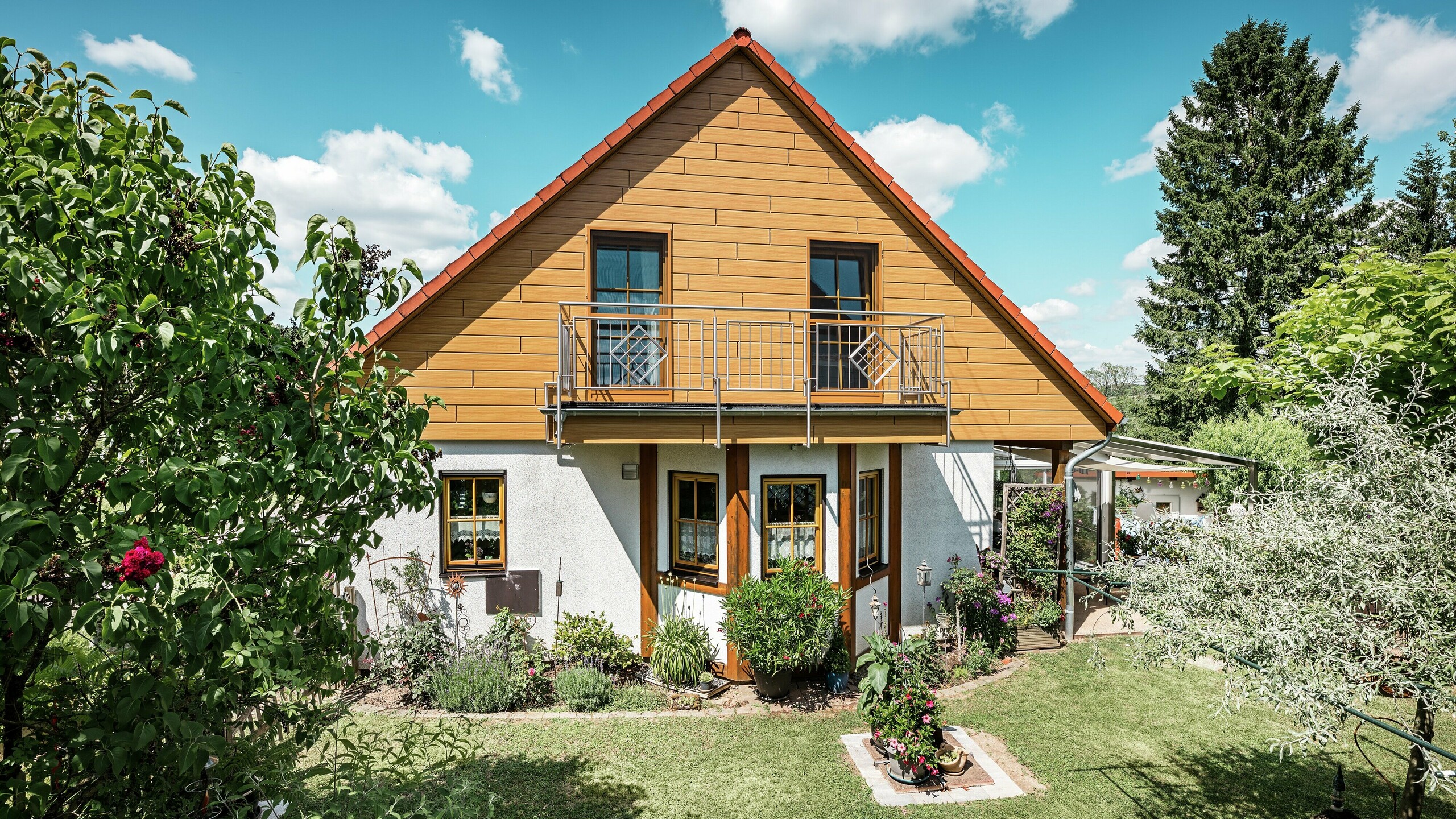 Auf diesem Bild strahlt die sanierte Giebelseite eines Einfamilienhauses in Blieskastel in warmem Eiche-Natur-Ton dank der PREFA Sidings. Die hochwertige Aluminiumfassade in realistischer Holzoptik setzt einen starken visuellen Akzent und harmoniert eindrucksvoll mit dem roten Ziegeldach und dem üppigen Garten. Umgeben von Blumen, Sträuchern und sattem Grün wirkt das Haus einladend und gepflegt. Die PREFA Oberfläche kombiniert natürliche Optik mit funktionalen Vorteilen wie hoher Witterungsbeständigkeit, Stabilität und nahezu wartungsfreier Langlebigkeit – ein ästhetisches Highlight mit nachhaltigem Mehrwert.