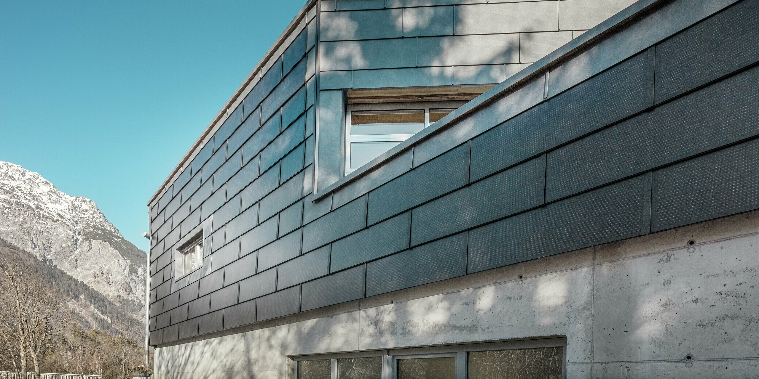 Fassade des Firmengebäudes der Spenglerei RÜMA in Vorarlberg mit innovativen PREFA Solardachplatten und Sichtbeton, umgeben von Bergen unter klarem blauem Himmel, mit Schatten von Bäumen auf der Wand.