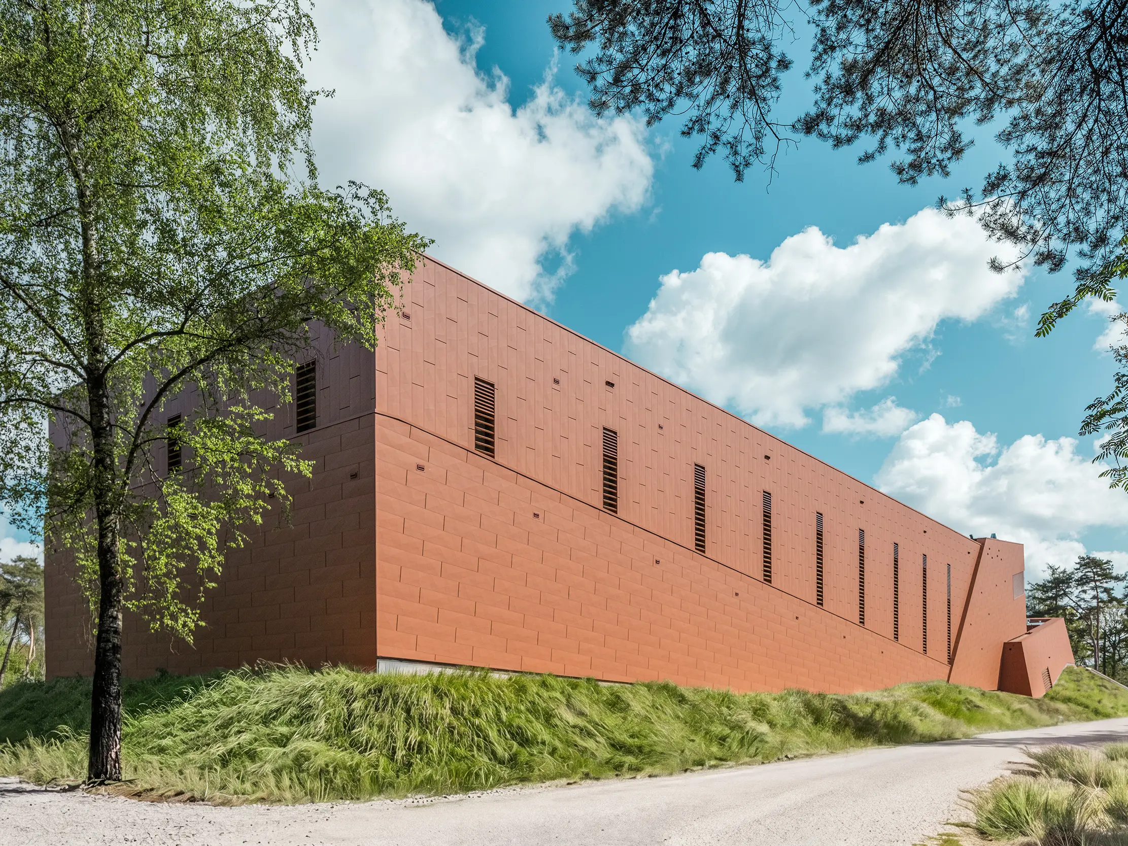 Das langgestreckte Trinkwasserpumpwerk WML in Lottum präsentiert sich als kraftvoller Baukörper inmitten einer naturnahen Landschaft aus Bäumen, Wiesen und Himmelspanorama. Die prägnante PREFA Siding.X Aluminiumfassade in warmer Rostoptik zieht als visueller Eyecatcher alle Blicke auf sich und fasziniert mit ihrer fein facettierten, lebendig strukturierten Oberfläche. Schlanke, vertikale Öffnungen gliedern die Fassade rhythmisch. Die langlebigen, leichten und recycelbaren PREFA Aluminium-Paneele bieten höchste Witterungsbeständigkeit, Farbkonstanz und eine nachhaltige, wartungsarme Gebäudehülle.