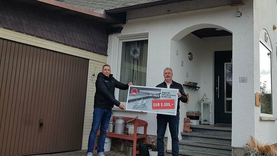 Zwei Männer stehen vor einem Haus und halten einen Scheck in der Hand.