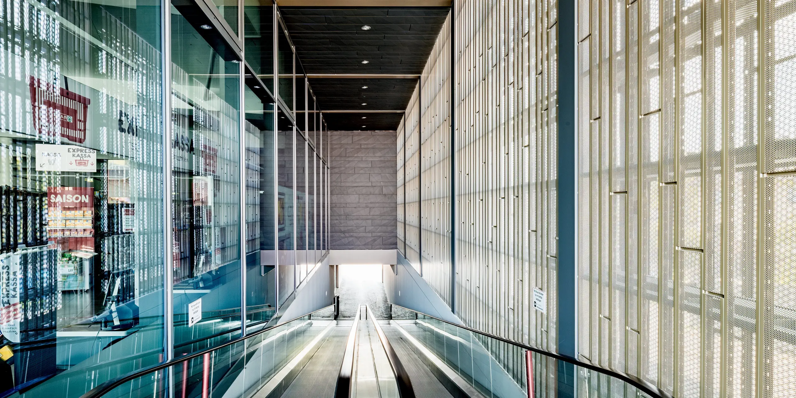 Das Bild zeigt den beeindruckenden Eingangsbereich des Einkaufszentrums in Rohrbach, der durch die Kombination moderner Materialien und funktionalem Design besticht. Im Vordergrund ist die Rolltreppe zu sehen, die die Besucher in die unteren Ebenen des Gebäudes führt. An den Wänden erkennt man die präzise angebrachten, perforierten Siding-Paneele in Anthrazit. Durch ihre leichte Textur und das spielerische Lichtspiel schaffen sie eine angenehme Atmosphäre. Diese Paneele sind nicht nur ästhetisch vorteilhaft, sondern sorgen auch für Belüftung und Lichtdurchlass und versorgen den Innenraum so optimal mit natürlichem Licht. Die großen Fensterflächen auf der linken Seite ermöglichen einen freien Blick nach außen und verstärken das Gefühl von Offenheit und Transparenz.