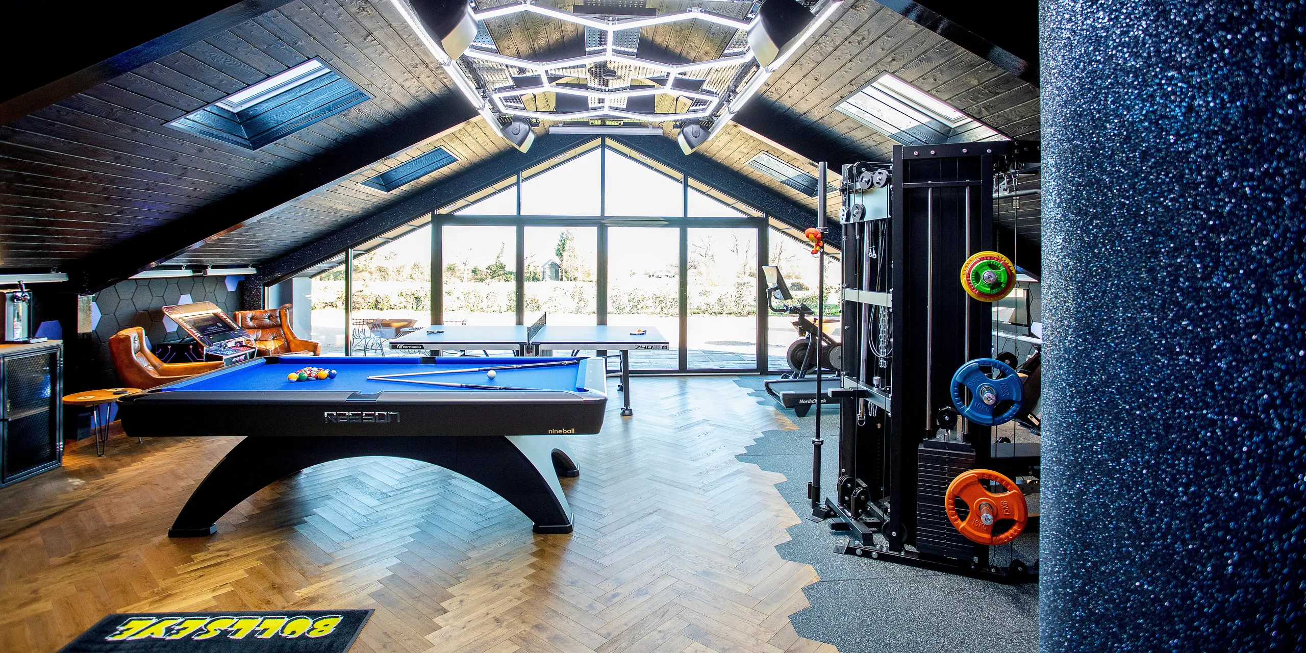 Innenansicht des modernen Freizeitgebäudes mit großflächiger Glasfront und markanter Dachkonstruktion. Der vielseitige Raum kombiniert Billardtisch, Trainingsgeräte, Tischtennistisch und Arcade‑Bereich. Hochwertige Materialien, dynamische Beleuchtung und Parkettboden schaffen eine einladende, energiegeladene Atmosphäre.