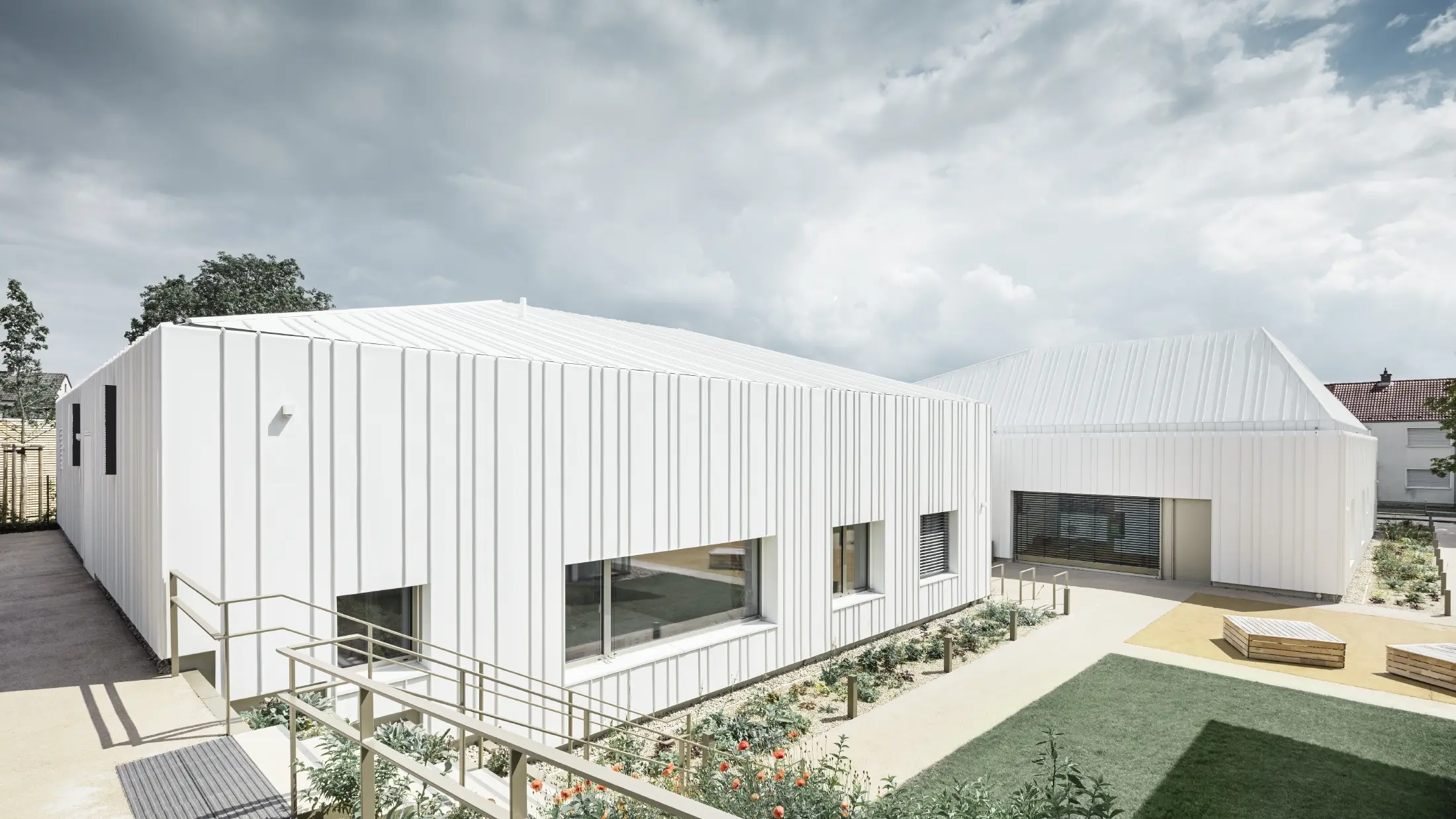 Modernes Kindergartengebäude mit klarer, geometrischer Architektur und Fassade aus weißen Aluminiumprofilen. Die Anlage besteht aus mehreren verbundenen Baukörpern mit flach geneigten Dächern. Große Fenster sorgen für viel Tageslicht, während der Außenbereich mit Wegen, Beeten und Sitzbänken gestaltet ist. Der Himmel ist teils bewölkt und verleiht der Szene eine ruhige Atmosphäre.