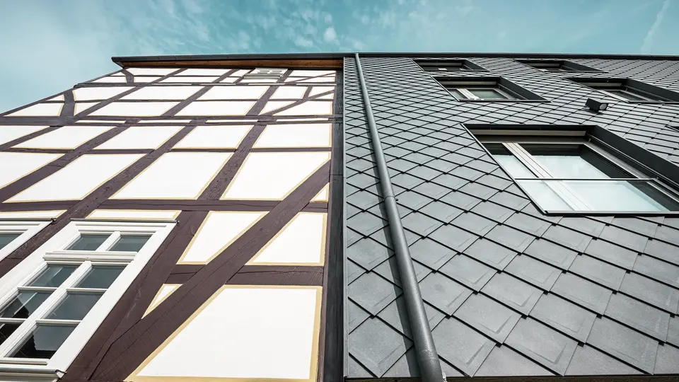 Nahaufnahme der Fassade des Volkshauses in Homberg, DE, mit modernen PREFA Wandrauten 29 × 29 in P.10 Steingrau und historischem Fachwerk. Die Aluminiumverkleidung vereint zeitlose Eleganz, Witterungsschutz und Langlebigkeit, harmonisch ergänzt durch eine integrierte Dachentwässerung. Die funktionelle Entwässerung des Regenwassers erfolgt über die PREFA Kastenrinne mit anschließendem Ablaufrohr. 