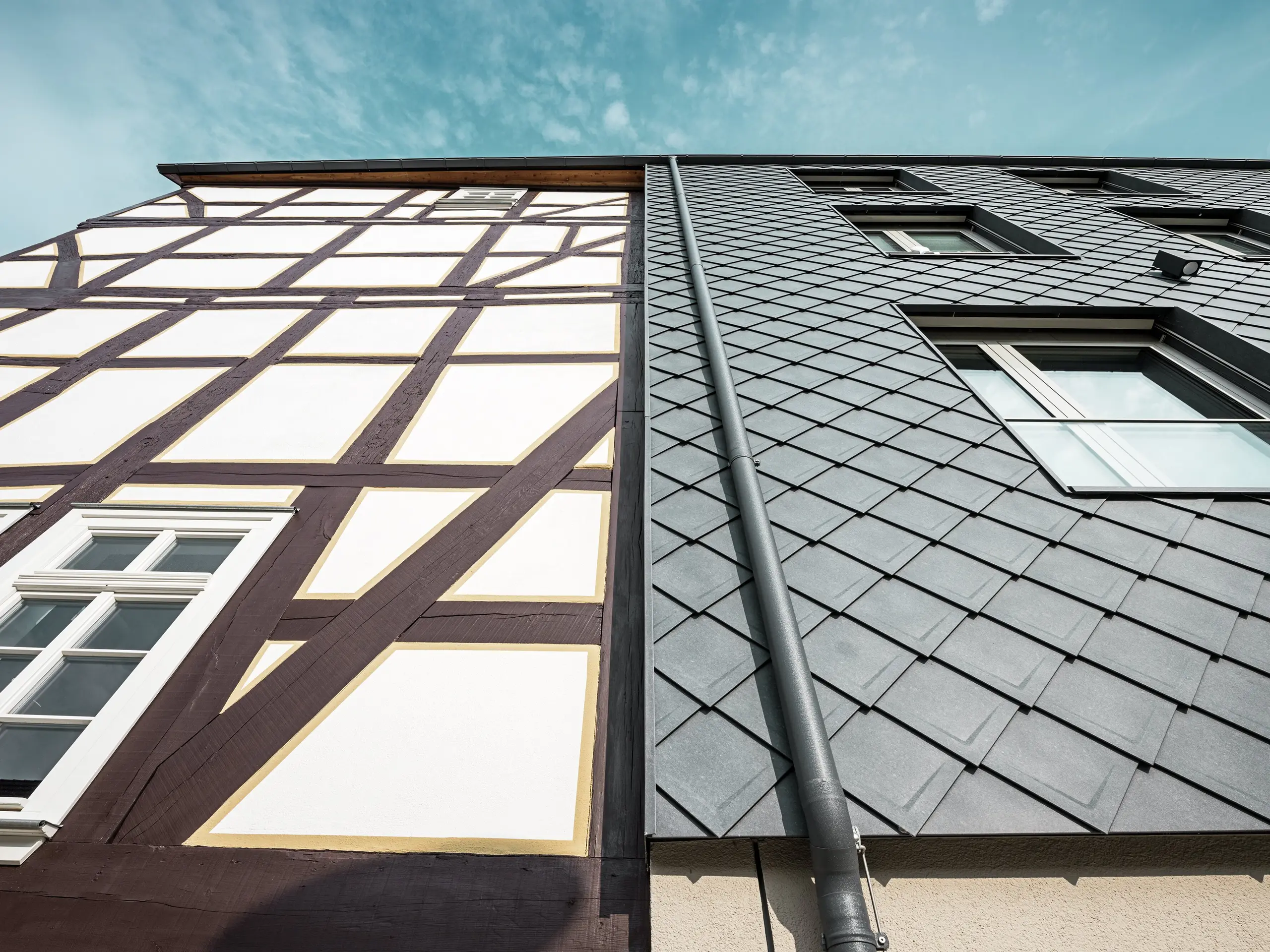 Nahaufnahme der Fassade des Volkshauses in Homberg, DE, mit modernen PREFA Wandrauten 29 × 29 in P.10 Steingrau und historischem Fachwerk. Die Aluminiumverkleidung vereint zeitlose Eleganz, Witterungsschutz und Langlebigkeit, harmonisch ergänzt durch eine integrierte Dachentwässerung. Die funktionelle Entwässerung des Regenwassers erfolgt über die PREFA Kastenrinne mit anschließendem Ablaufrohr. 