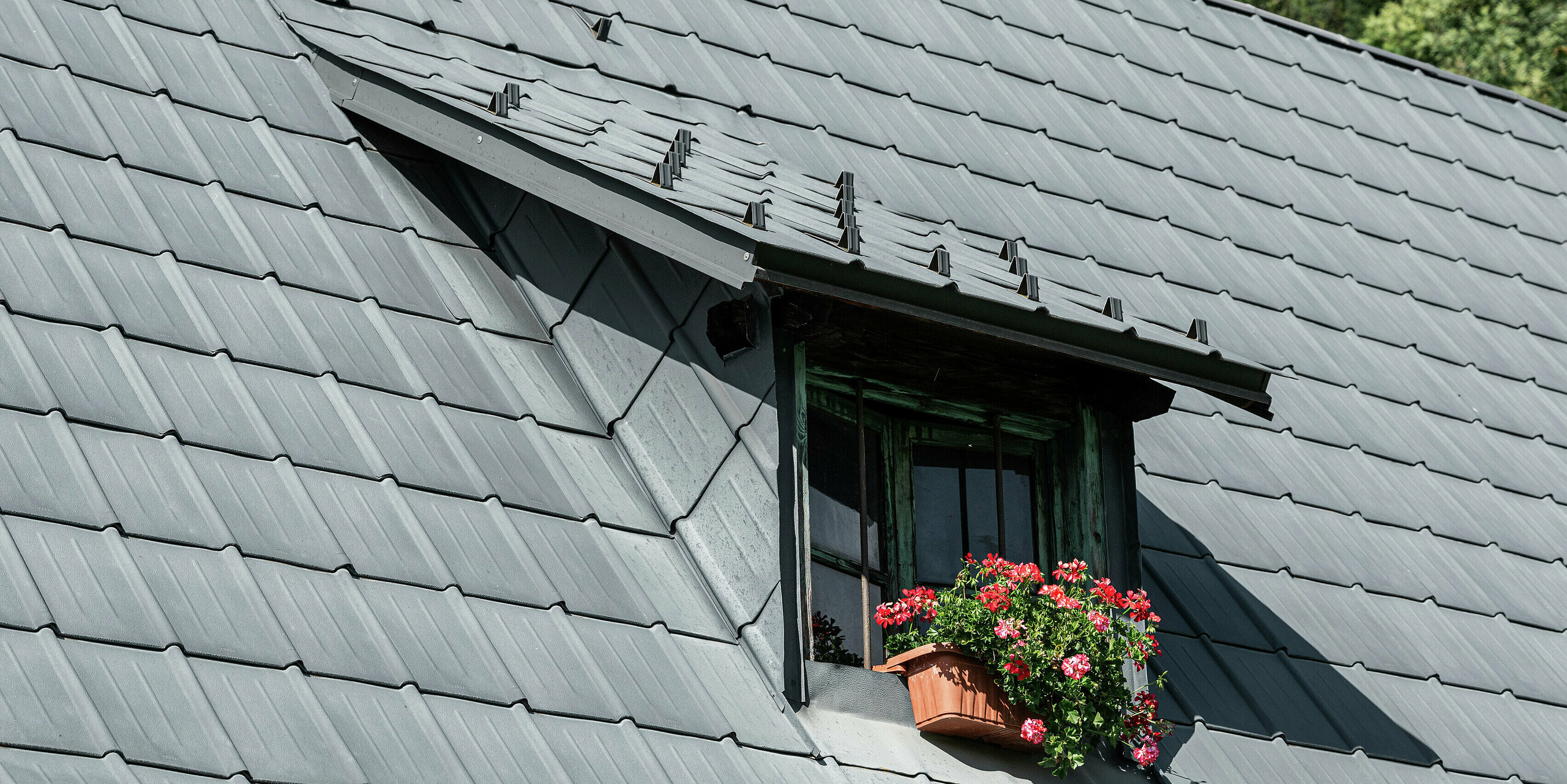 Das Einfamilienhaus im italienischen Tarvis, seit 1972 mit langlebigen PREFA Dachplatten eingedeckt, besticht durch seine markante Schleppgaube in der Mitte des Krüppelwalmdachs. Diese Gaube fügt sich harmonisch in das großzügige Aluminiumdach ein und sorgt für zusätzlichen Lichteinfall im Dachgeschoss. Die widerstandsfähigen Dachplatten schützen das Haus seit Jahrzehnten zuverlässig vor alpinen Witterungseinflüssen. Die mit Blumen geschmückte Gaube setzt einen reizvollen Akzent und unterstreicht die gelungene Verbindung von Funktionalität und Ästhetik.