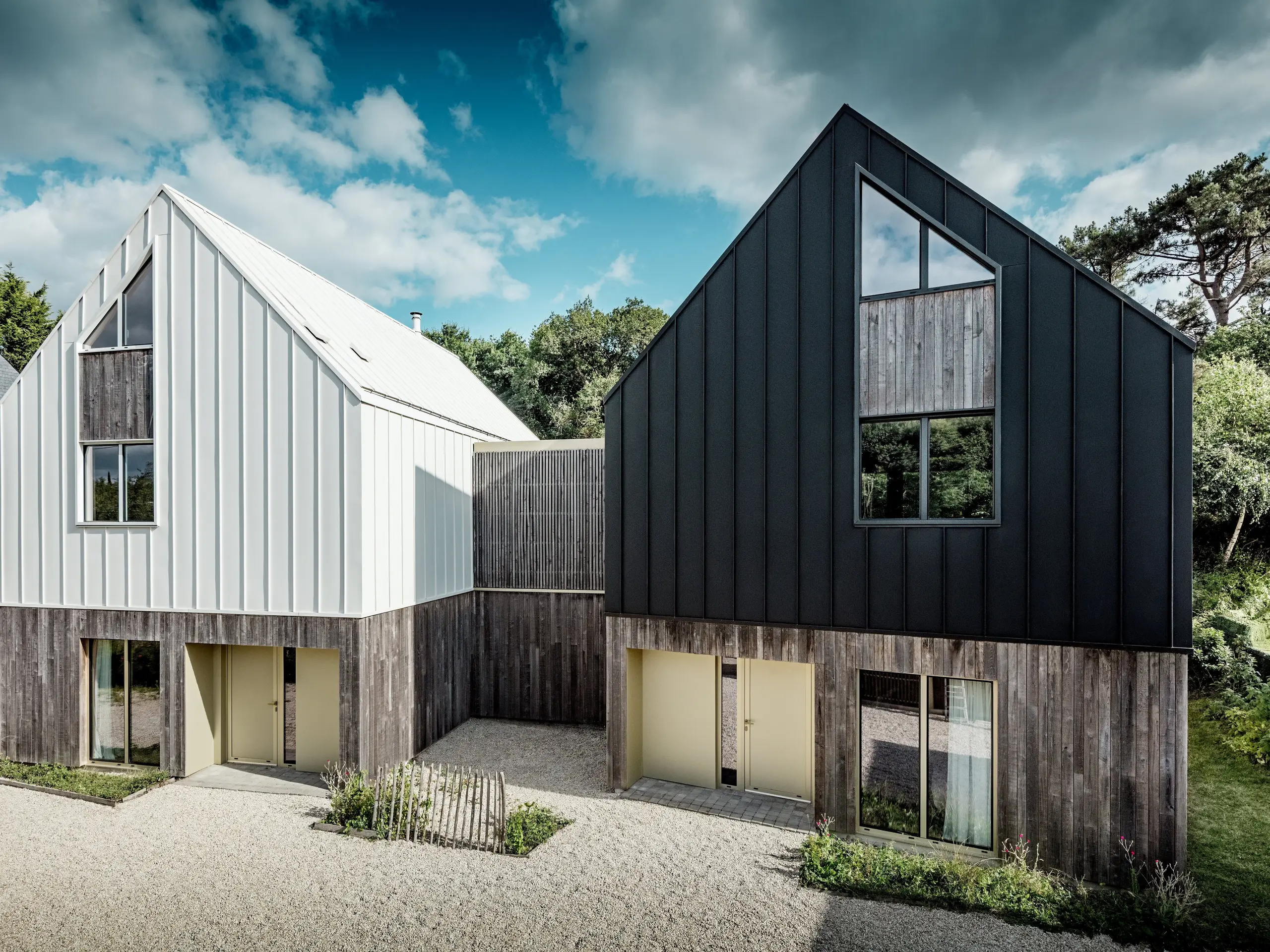 Die Frontansicht des Wohnensembles Le Rhune zeigt zwei moderne Baukörper mit markanten Satteldächern und kontrastierenden PREFALZ Aluminiumhüllen in Reinweiß und Schwarzgrau. Vor der Kulisse eines dramatischen Wolkenhimmels treten die klaren Linien der durchgängigen Stehfalzfassaden besonders eindrucksvoll hervor. Inspiriert von traditionellen bretonischen Fischerhäusern vereint die Architektur regionale Bautradition mit zeitgemäßem Design. Die langlebige und wetterbeständige PREFALZ Gebäudehülle verbindet Dach und Fassade nahtlos miteinander, ermöglicht präzise Detailausbildungen und verleiht dem Ensemble eine moderne, hochwertige sowie dauerhaft attraktive Erscheinung.