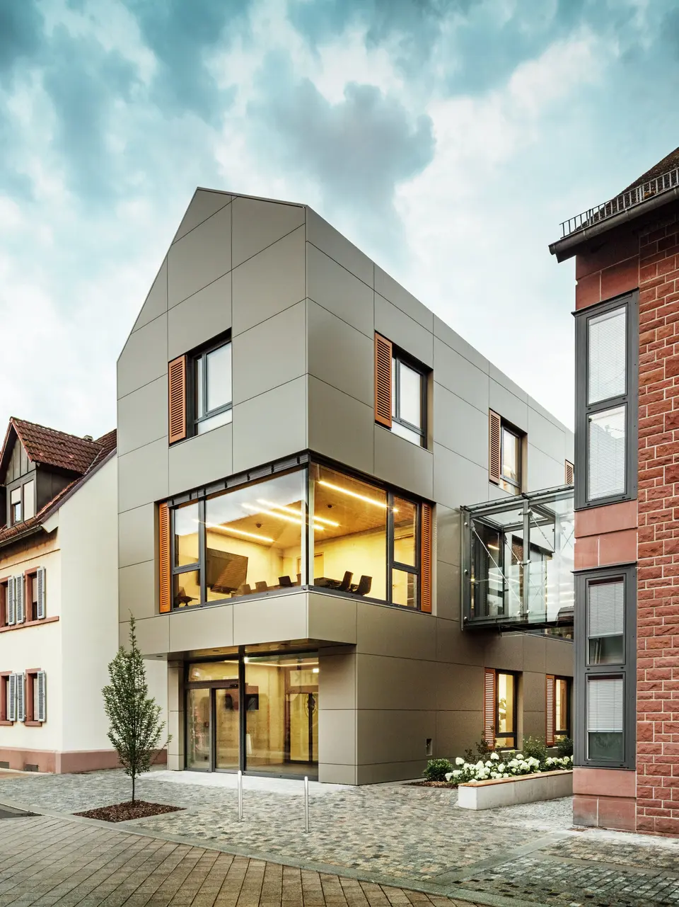 Das Bild zeigt das moderne Rathaus in Alzenau, dessen Fassade mit PREFA-Aluminiumverbundplatten in einem warmen Bronzeton verkleidet ist. Große Glasflächen unterbrechen die Fassade und geben den Blick auf ein lichtdurchflutetes Interieur frei. Ein gläserner Verbindungsgang auf der rechten Bildseite verbindet das Gebäude mit einem nahegelegenen traditionellen Backsteingebäude und schafft so einen interessanten Kontrast zwischen moderner und historischer Architektur.