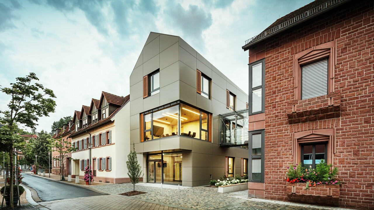 Das Bild zeigt das moderne Rathaus in Alzenau, dessen Fassade mit PREFA-Aluminiumverbundplatten in einem warmen Bronzeton verkleidet ist. Große Glasflächen unterbrechen die Fassade und geben den Blick auf ein lichtdurchflutetes Interieur frei. Ein gläserner Verbindungsgang auf der rechten Bildseite verbindet das Gebäude mit einem nahegelegenen traditionellen Backsteingebäude und schafft so einen interessanten Kontrast zwischen moderner und historischer Architektur.